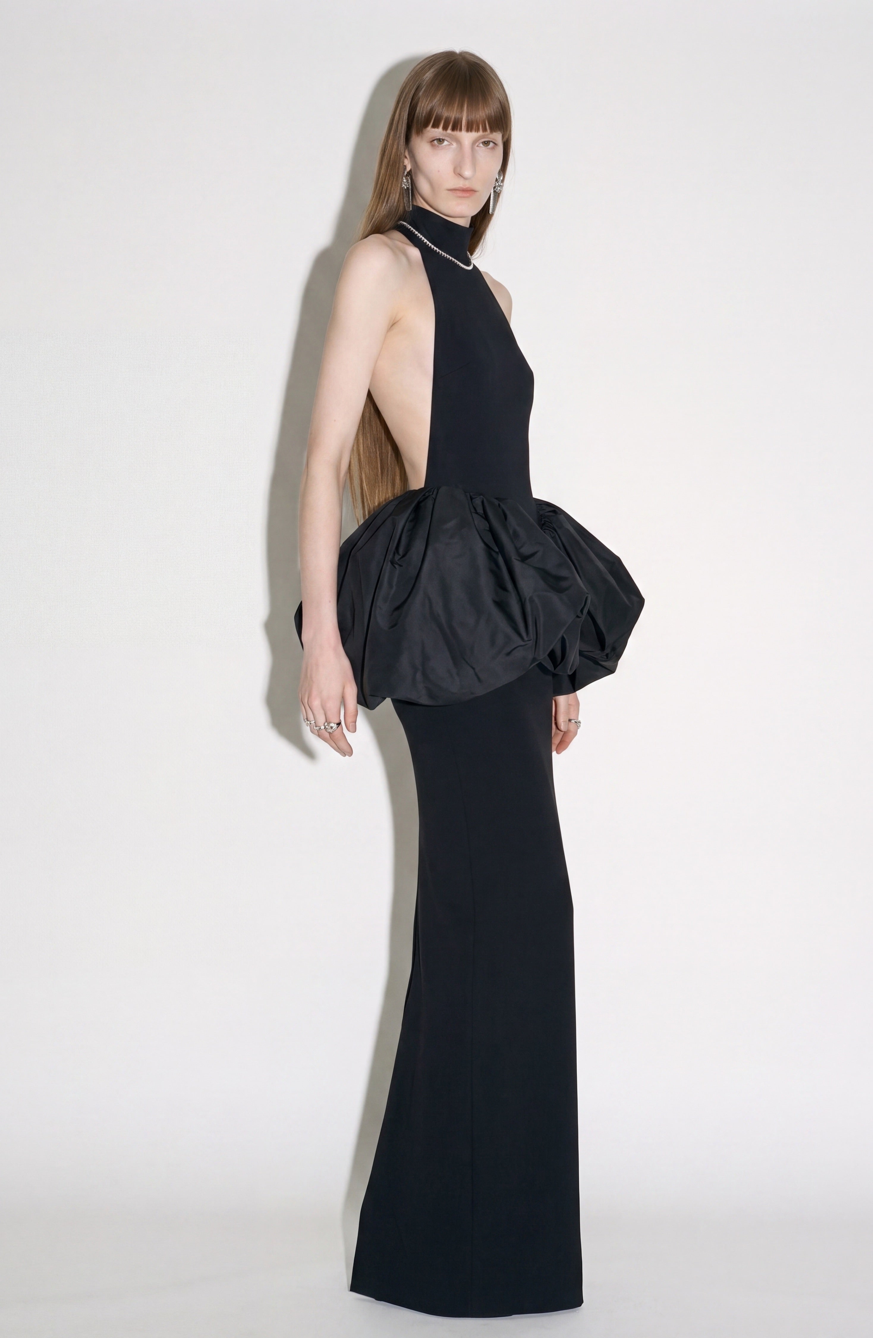 A person wearing the LOULOU Peplum Maxi Dress—a sleeveless, backless black evening gown featuring a voluminous peplum detail at the hips—stands against a plain light background, looking toward the camera.