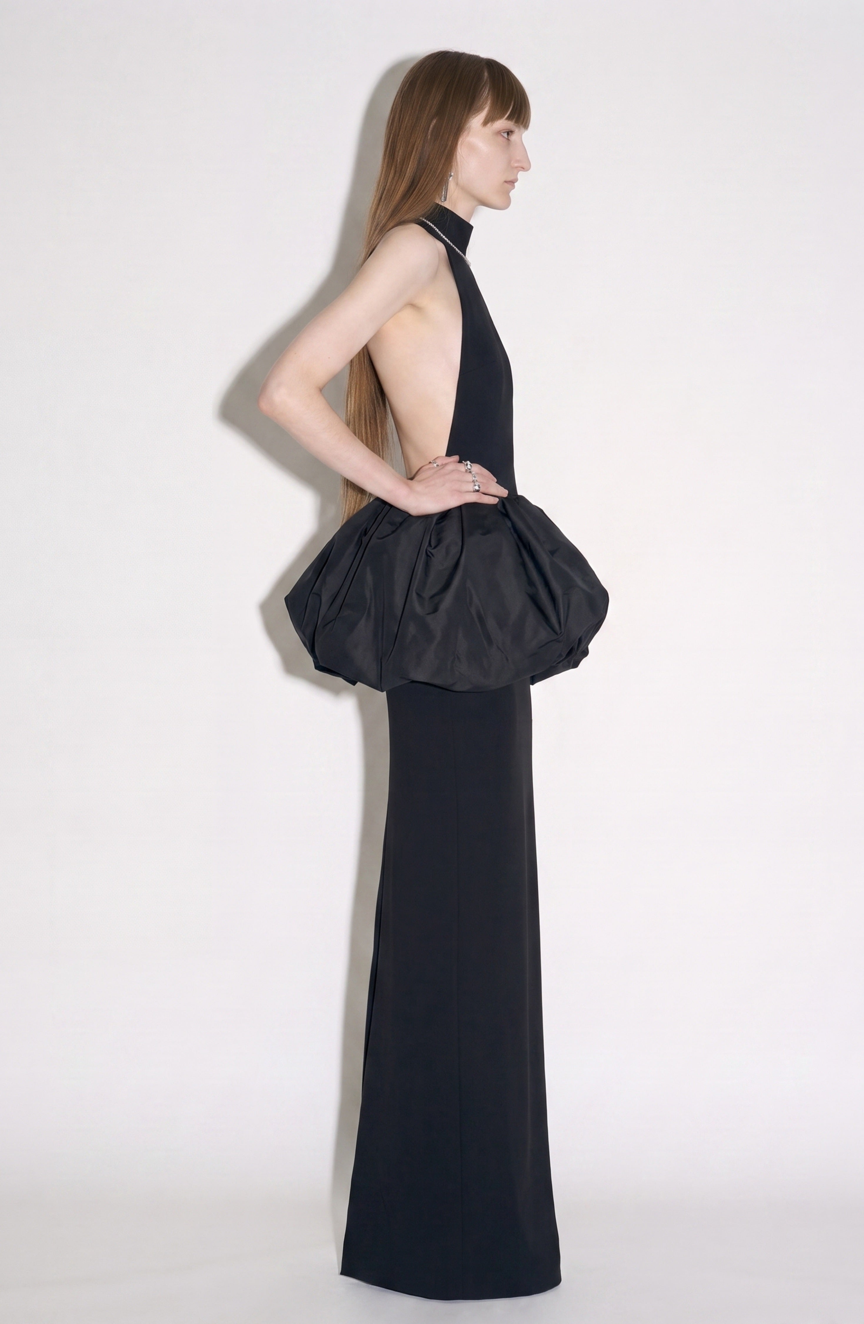 A person stands in profile wearing the LOULOU Peplum Maxi Dress, a sleeveless black evening gown featuring a high neckline, voluminous structured peplum at the hips, an open back, and a long, straight silhouette. The background is plain white.