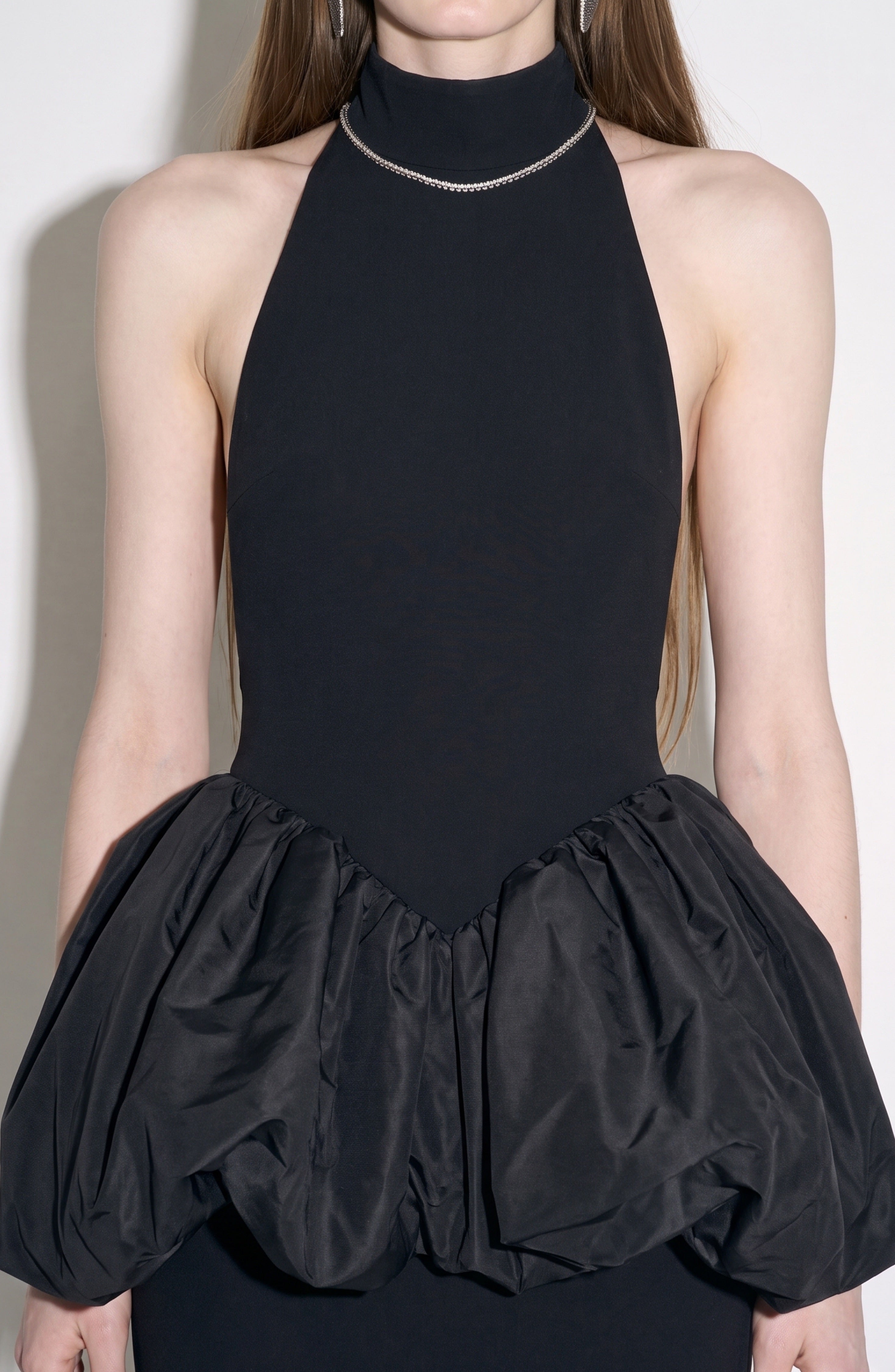 A person wearing the LOULOU Peplum Maxi Dress—a sleeveless black halter evening gown featuring a high neckline and a voluminous bubble peplum skirt—styled with a simple chain necklace. The photo is cropped from the neck down to the hips.