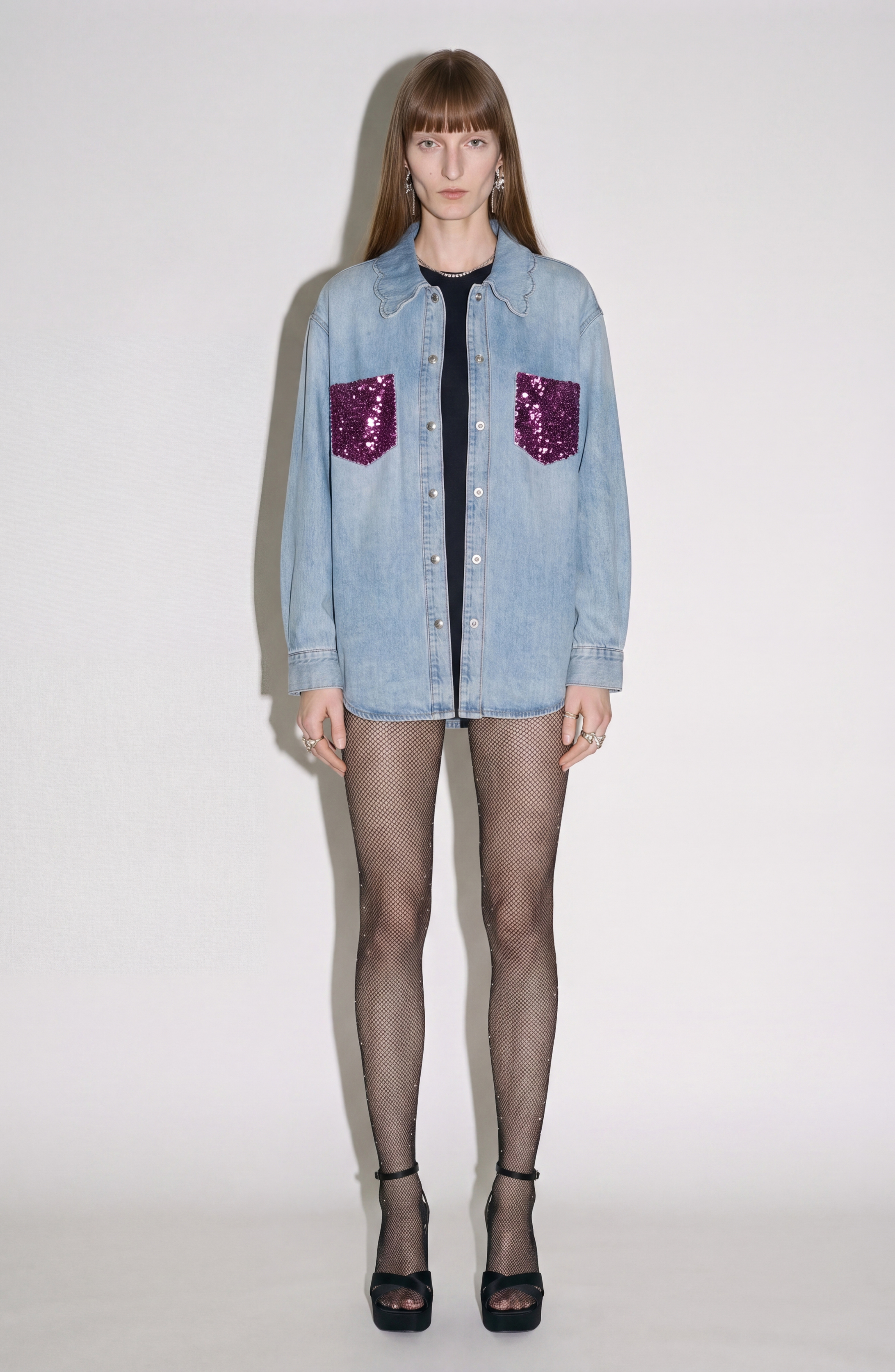 A person stands against a plain background wearing the LOULOU Sequin Pocket Denim Shirt, which features sequin chest pockets and a scalloped collar, paired with black fishnet tights and black heels.