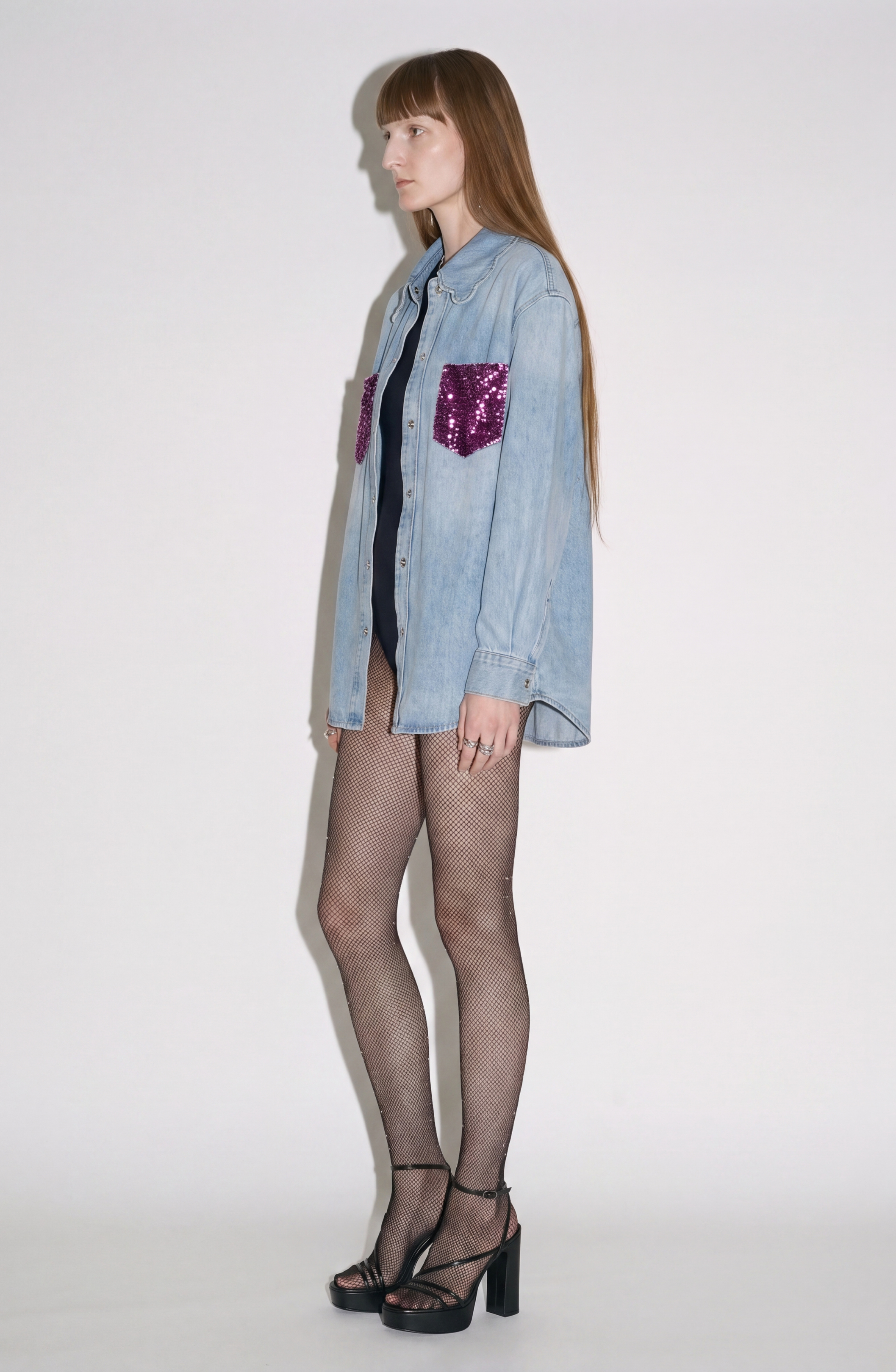A woman stands in profile against a plain white background wearing the LOULOU Sequin Pocket Denim Shirt—a light wash denim top featuring a scalloped collar and purple sequin chest pockets—paired with a black bodysuit, fishnet tights, and black platform heels.