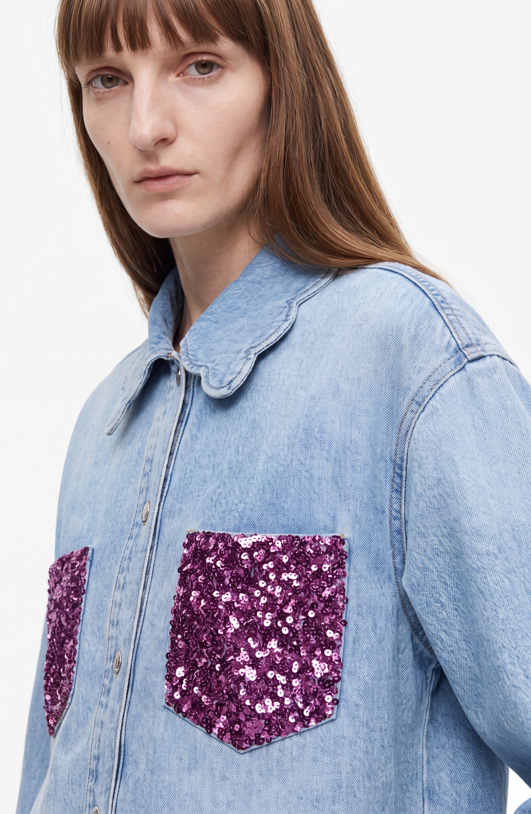 A person wearing the LOULOU Sequin Pocket Denim Shirt—a light wash denim shirt featuring a scalloped collar and two chest pockets adorned with shiny purple sequins—looks at the camera with a neutral expression.