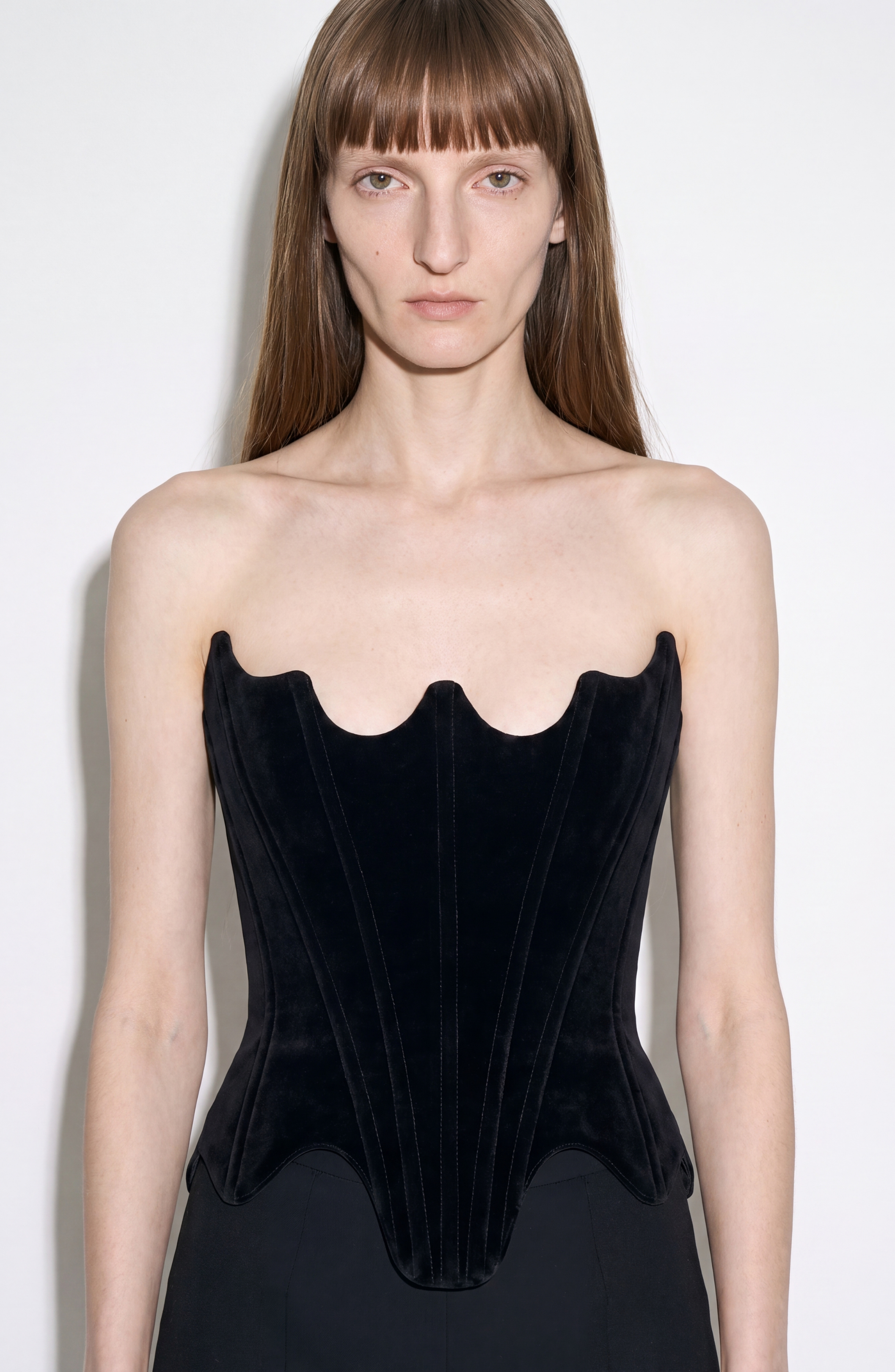 A person with long brown hair models the LOULOU Velvet Architectural Corset, a strapless black velvet piece with an architectural neckline and sculpted top edge, set against a plain white background.