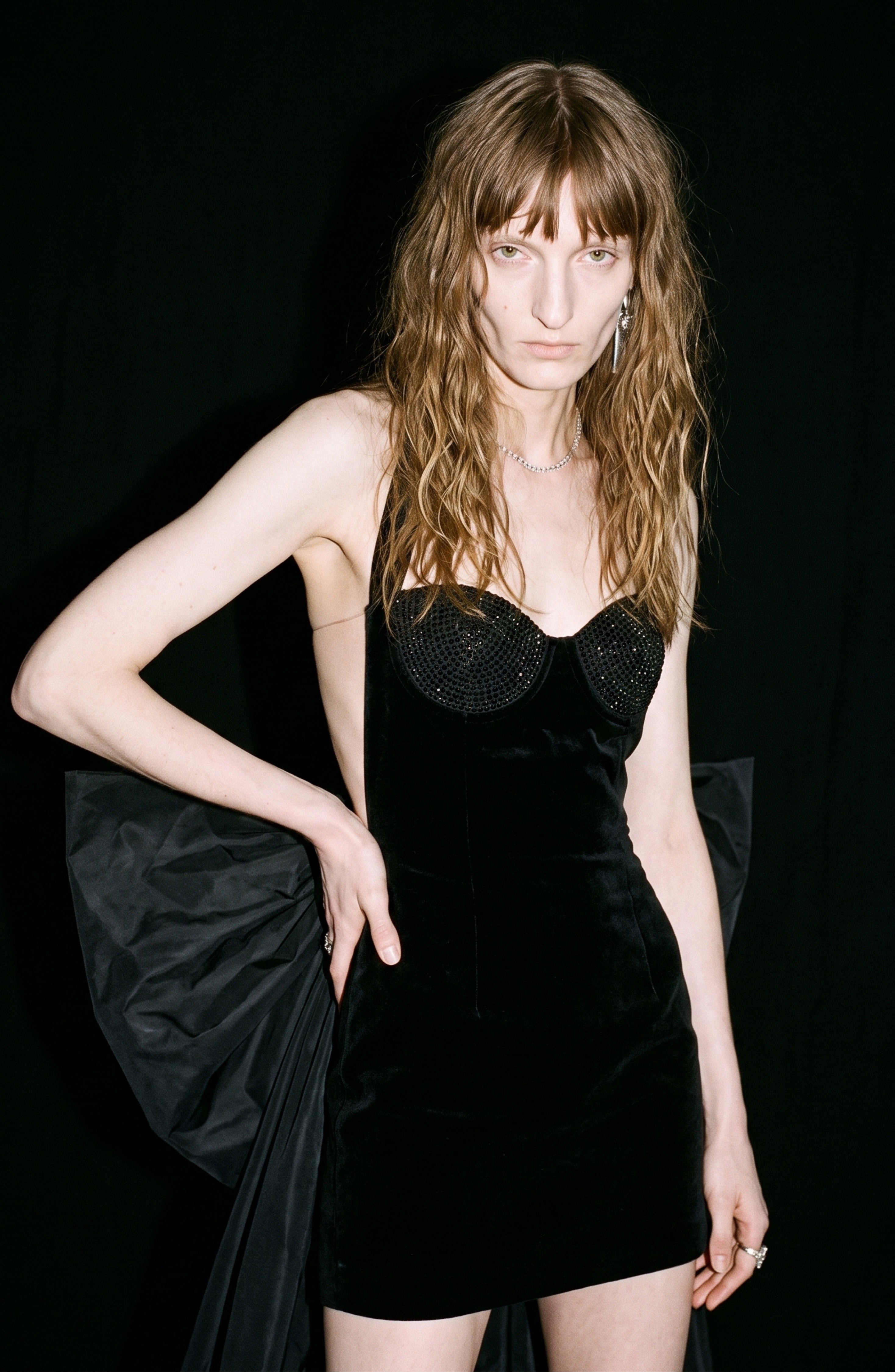 A person with long wavy hair wears the LOULOU Velvet Halter Bow Mini Dress, featuring a dramatic oversized bow in back, standing against a black background.