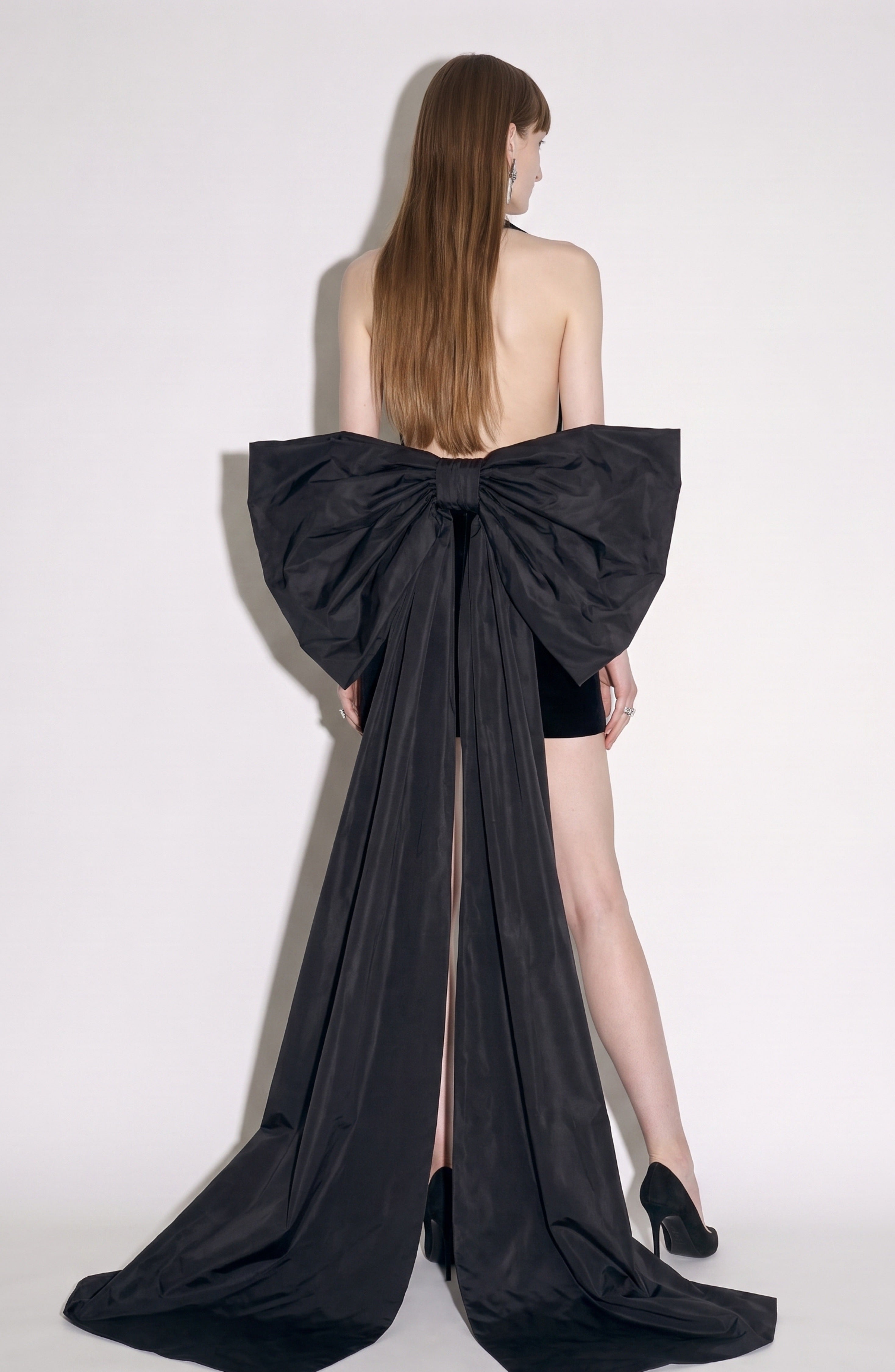 A woman models the LOULOU Velvet Halter Bow Mini Dress, standing against a plain white background. The dress features a luxurious black velvet fabric and a striking oversized bow with long tails attached at the lower back. She has long, straight brown hair and completes the look with black heels.