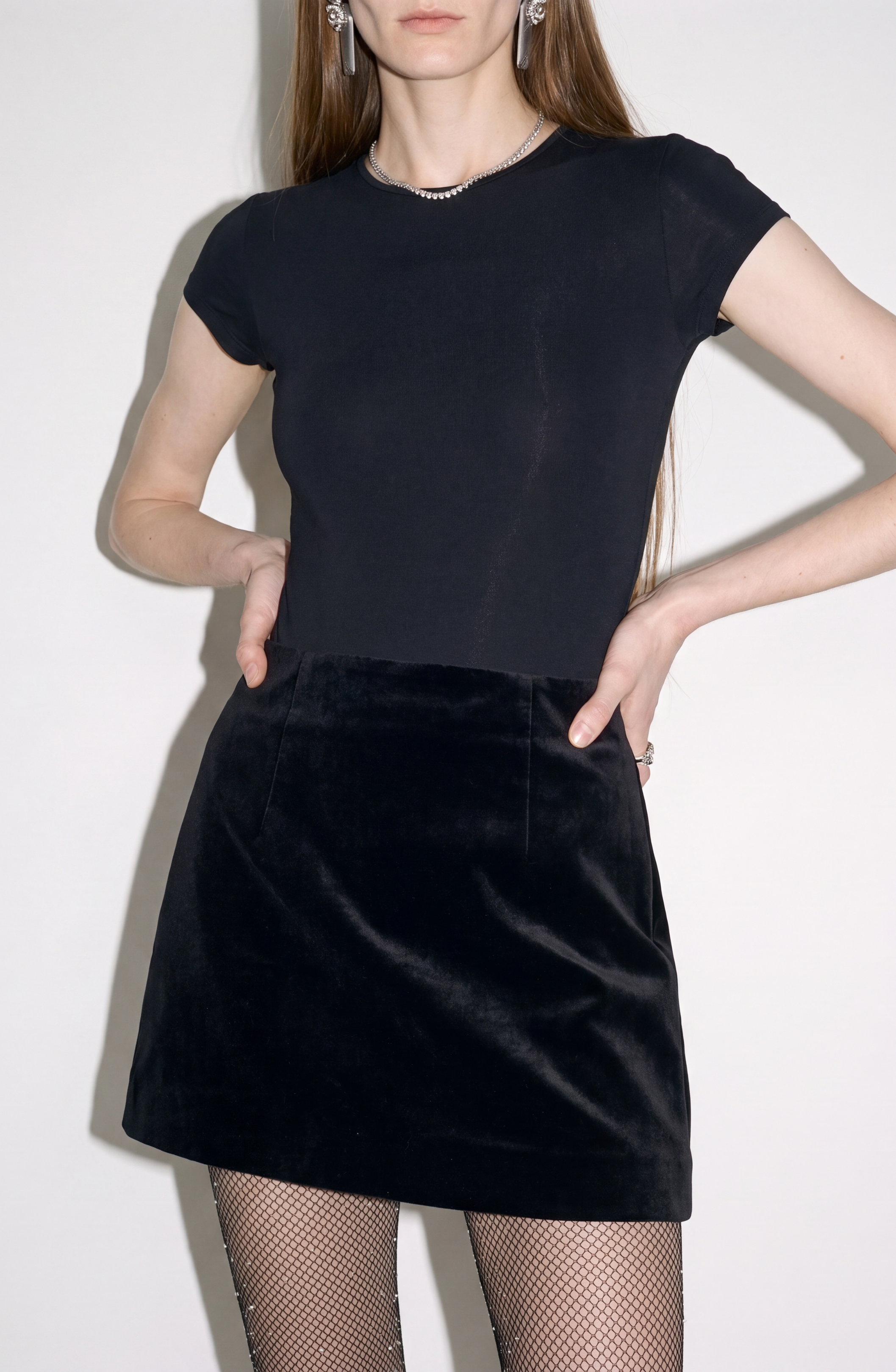 A woman stands against a plain light background, wearing a short-sleeved black top, fishnet tights, and the LOULOU Velvet Mini Skirt—a high-waisted black velvet mini skirt. Her long hair is down, and her hands rest on her hips.