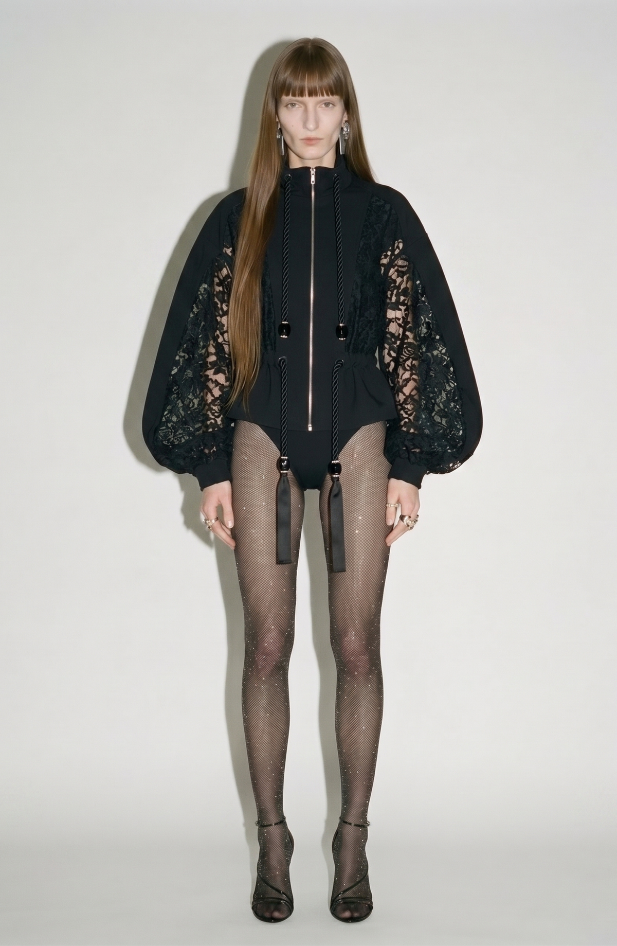 A model faces forward, wearing the LOULOU After Hours Bomber with floral-lace sleeves, a leotard, sparkly fishnet tights, and strappy black heels.