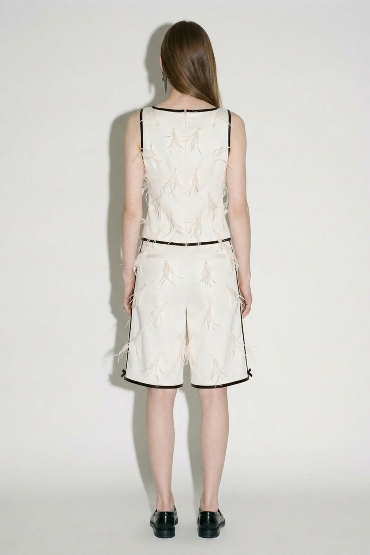 A woman with straight brown hair faces a white wall, wearing LOULOU's Applique Bermuda Shorts—ivory with subtle patterns and black trim—a sleeveless cream top, and black shoes. Her arms rest by her sides.