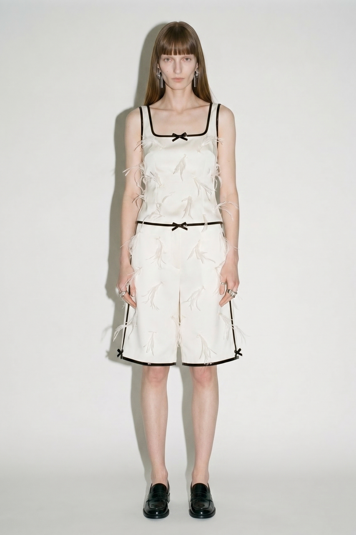 A woman with long, straight hair wears LOULOU's Applique Bermuda Shorts—ivory with black trim, bows, and feather accents—paired with a matching sleeveless top and classic black loafers against a plain white background.