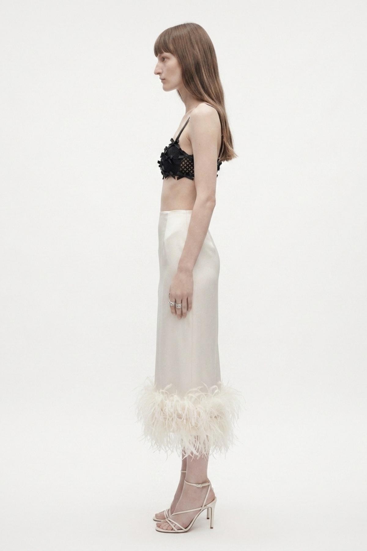 A woman stands in profile against a white background, wearing the LOULOU Black Bow Appliqué Bralette Top, a cream satin skirt with feather trim at the hem, and strappy high-heeled sandals.