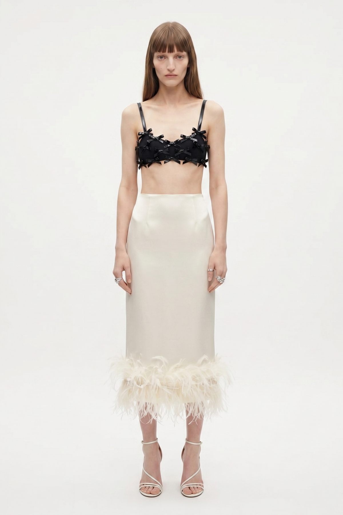 A woman with straight brown hair and a neutral expression wears the LOULOU Black Bow Appliqué Bralette Top against a plain white background, styled with a high-waisted ivory satin feather-hem midi skirt.