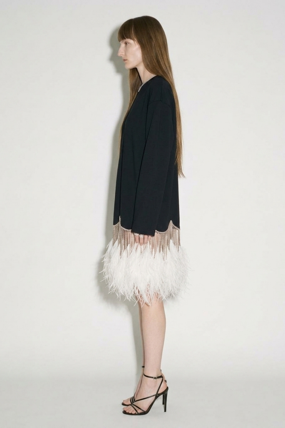 A woman with long brown hair stands in profile against a plain white background, wearing the LOULOU Black Crystal Feather Long Sleeve Mini Dress paired with black strappy heels.