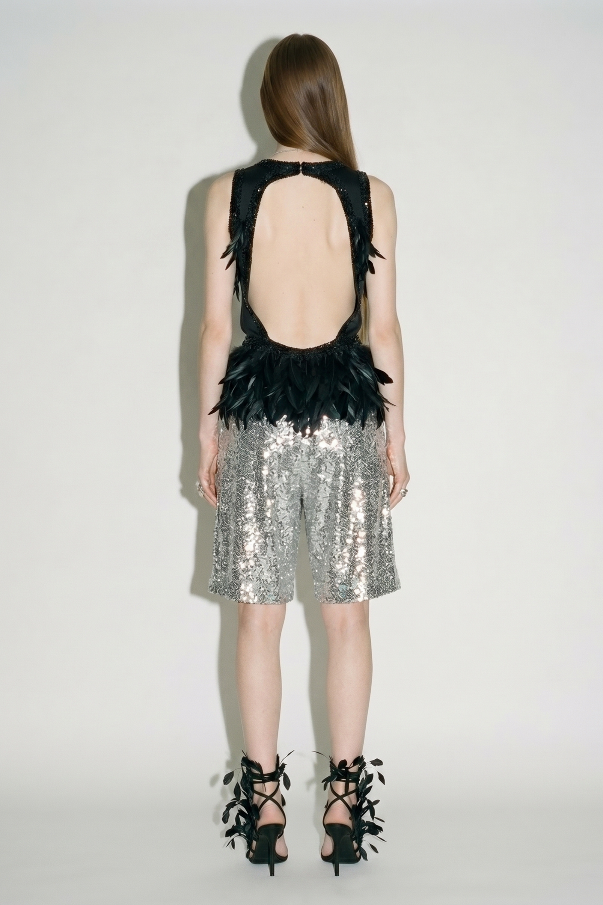 Back view of the LOULOU Black Feather Sequin Peplum Top, styled with silver sequined shorts and black ribbon-and-leaf heels.