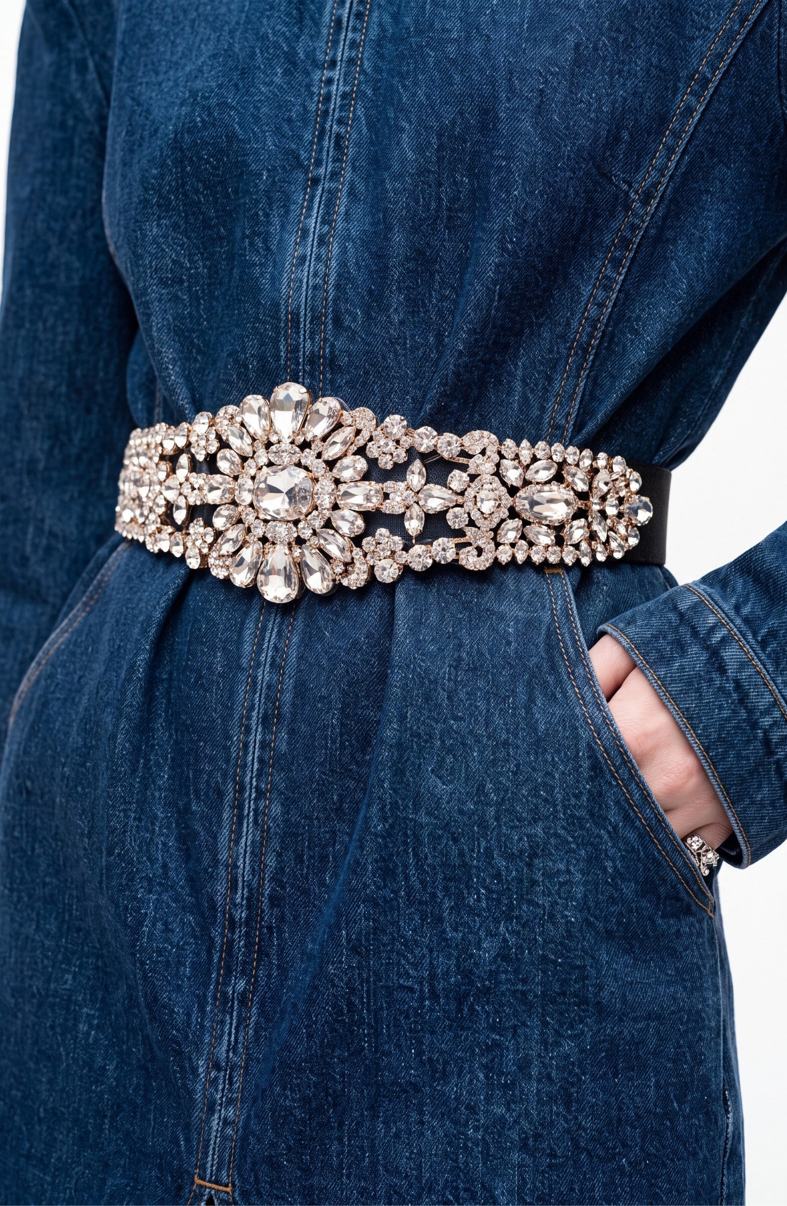 A person wears the LOULOU Crystal Belt Denim Dress—a dark indigo midi dress featuring a wide, ornate belt dazzling with large and small crystal embellishments—one hand tucked into the pocket, drawing attention to the sparkling gemstone-adorned accessory.