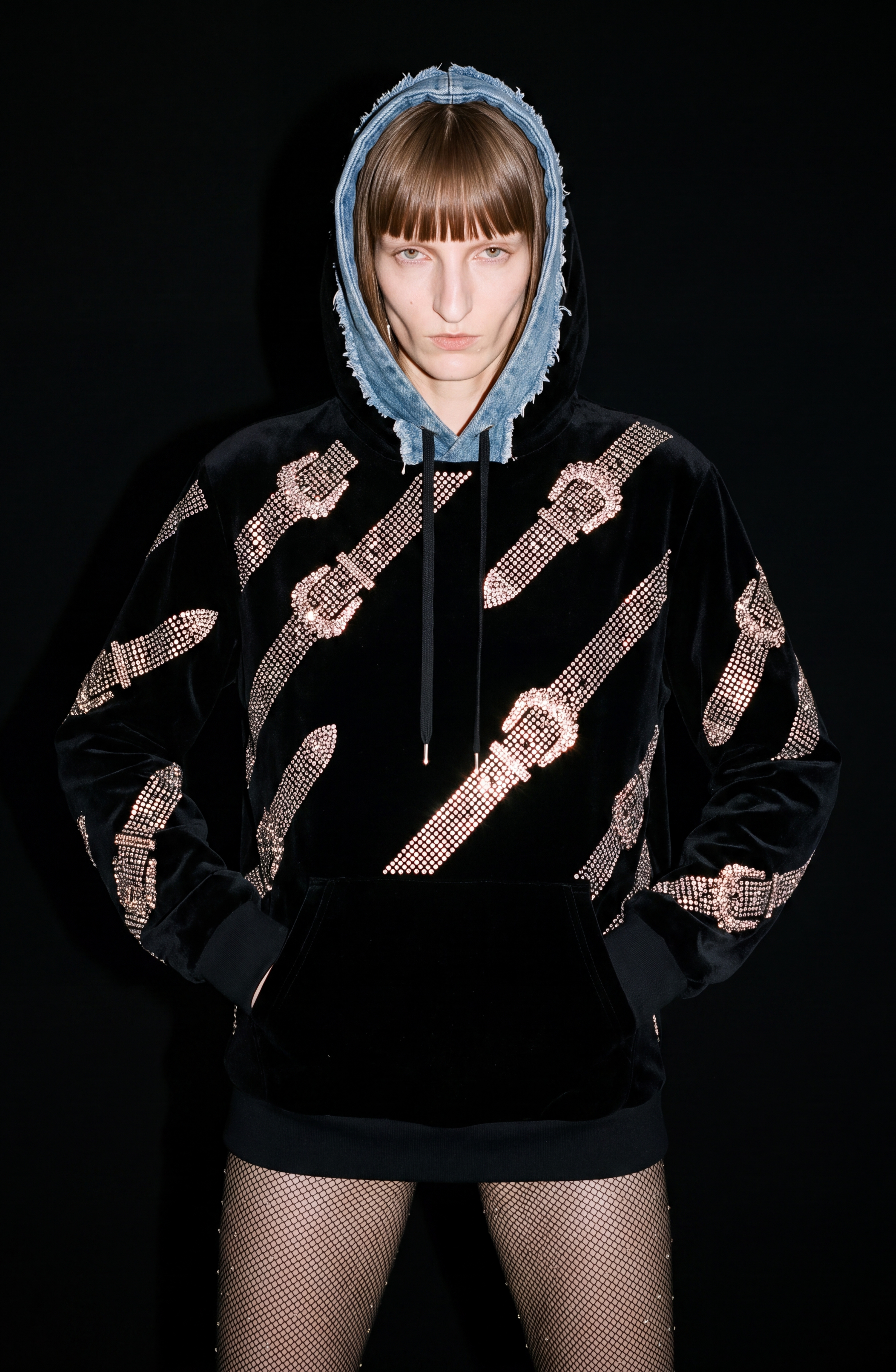 A person with straight hair wears the LOULOU Crystal Belt Velvet Hoodie, featuring sparkling belt buckle details and a blue denim hood, paired with fishnet stockings against a black backdrop.