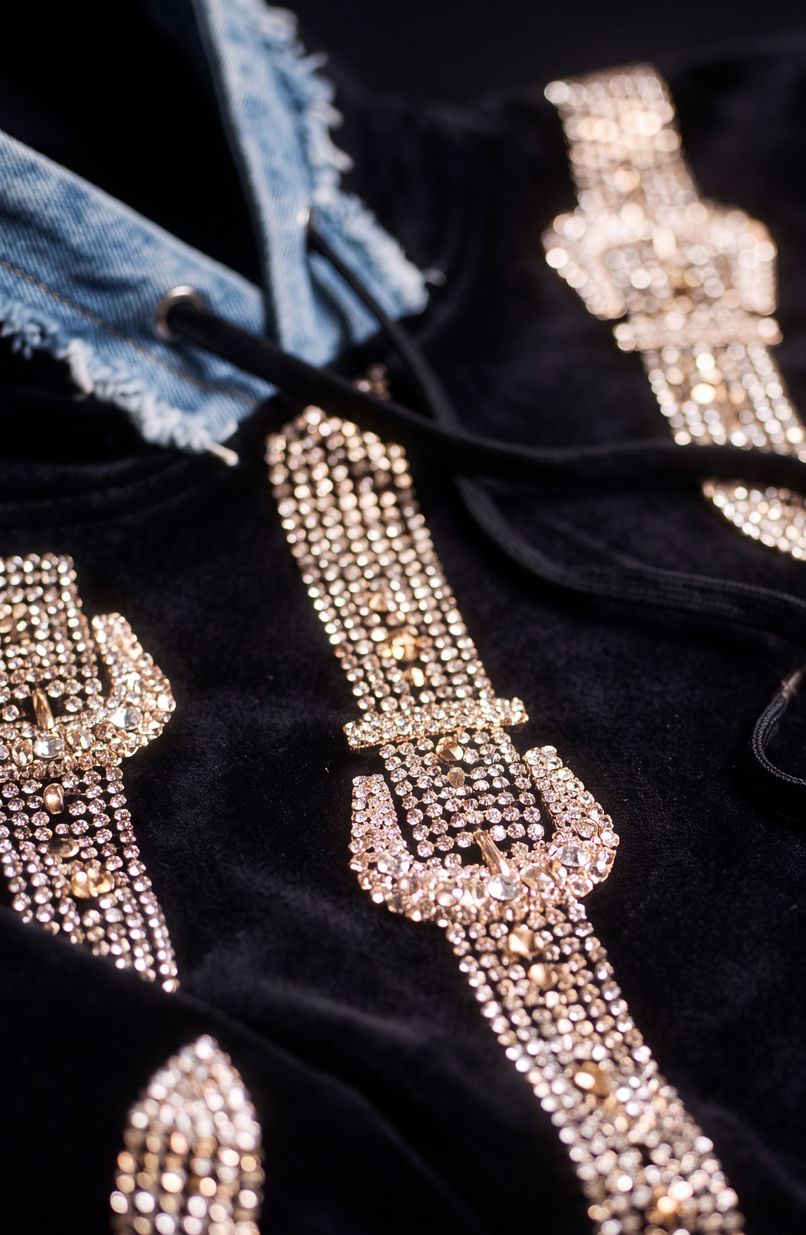 A close-up of the LOULOU Crystal Belt Velvet Hoodie, showcasing its black velvet fabric adorned with sparkling gold and silver studs in intricate patterns, paired with a blue denim hood featuring frayed edges and black drawstrings.