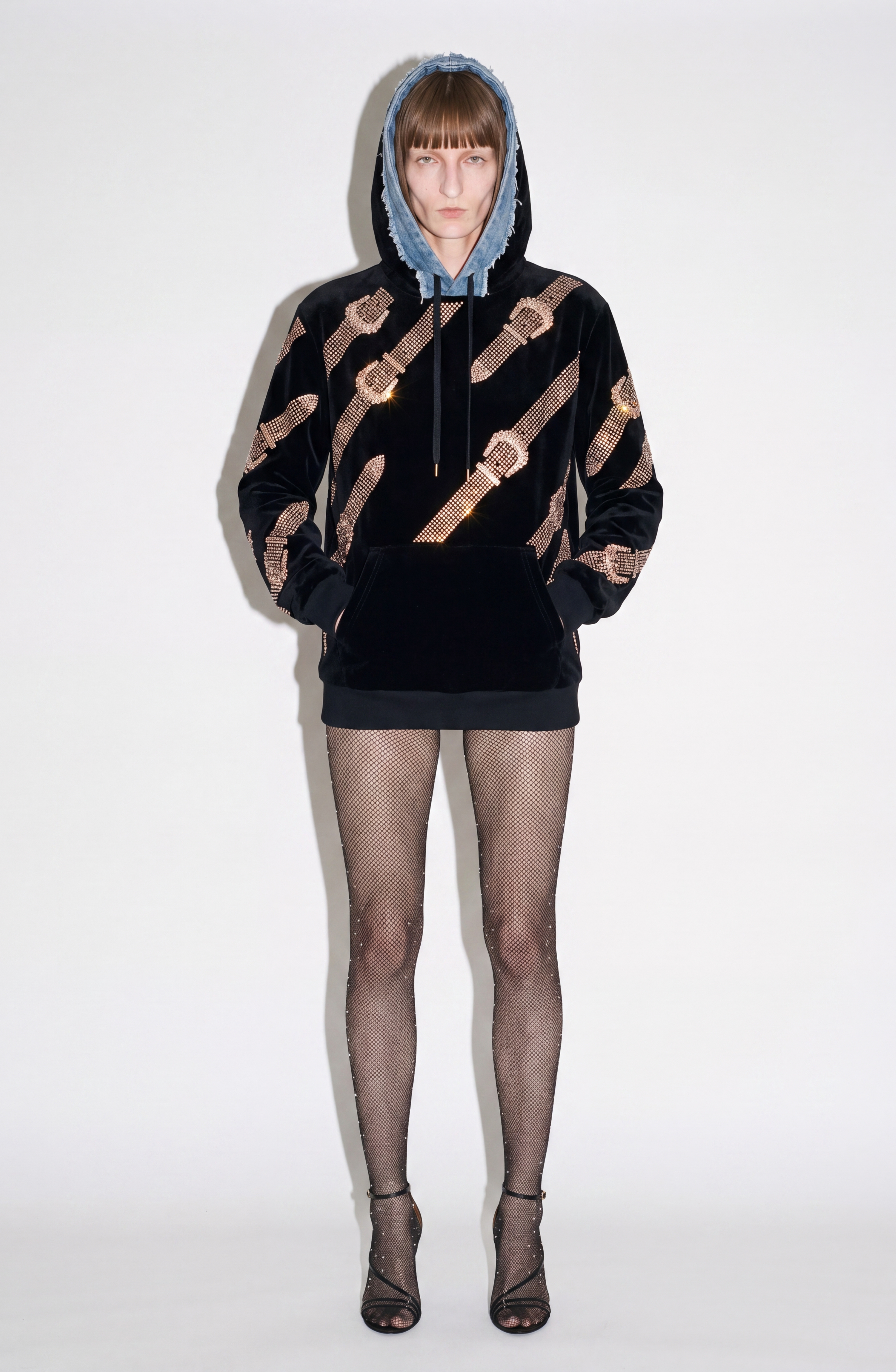 A person stands facing forward against a plain white background, wearing the LOULOU Crystal Belt Velvet Hoodie in black with gold diagonal stripes and a blue hood, paired with fishnet stockings and black high-heeled sandals.