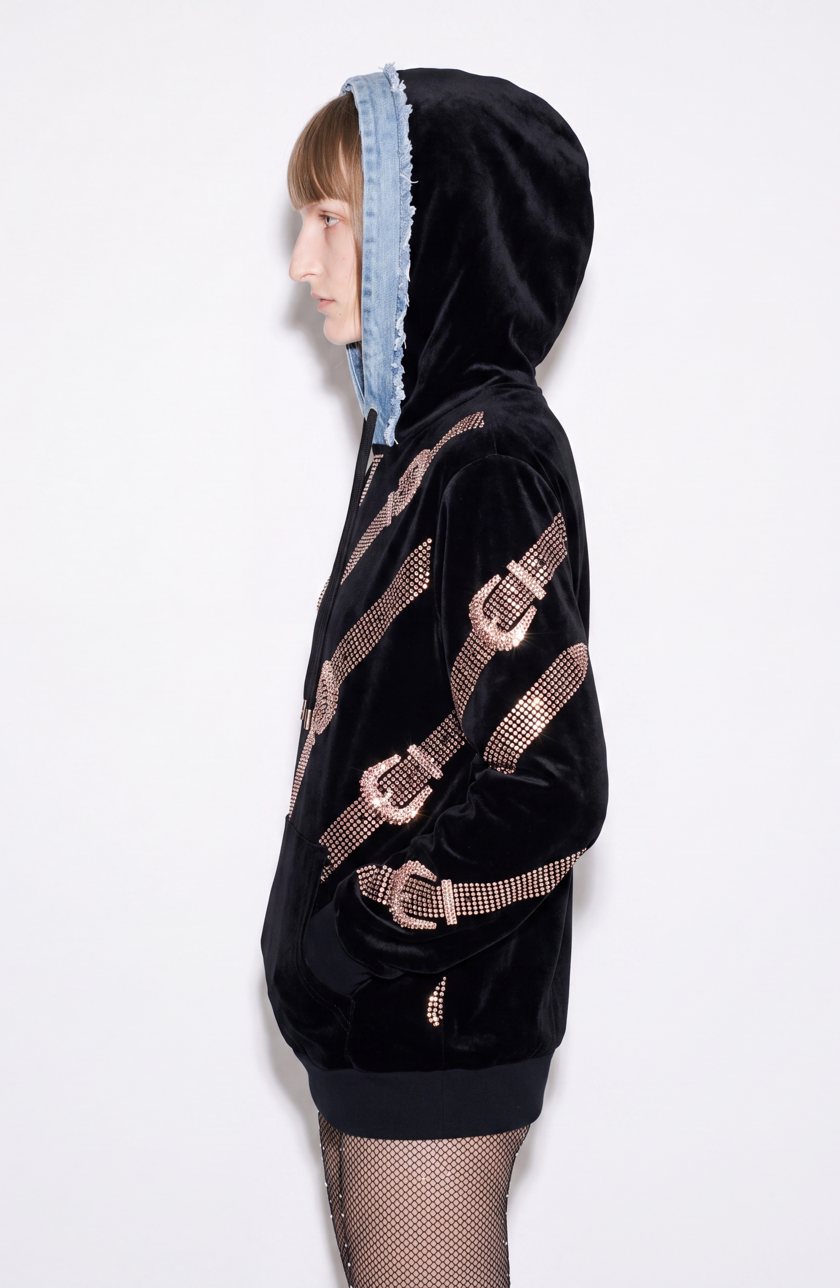 A person in profile wears the LOULOU Crystal Belt Velvet Hoodie—a black velvet hoodie with shiny pink diagonal stripes and a frayed denim hood—paired with fishnet tights, standing against a plain white background.