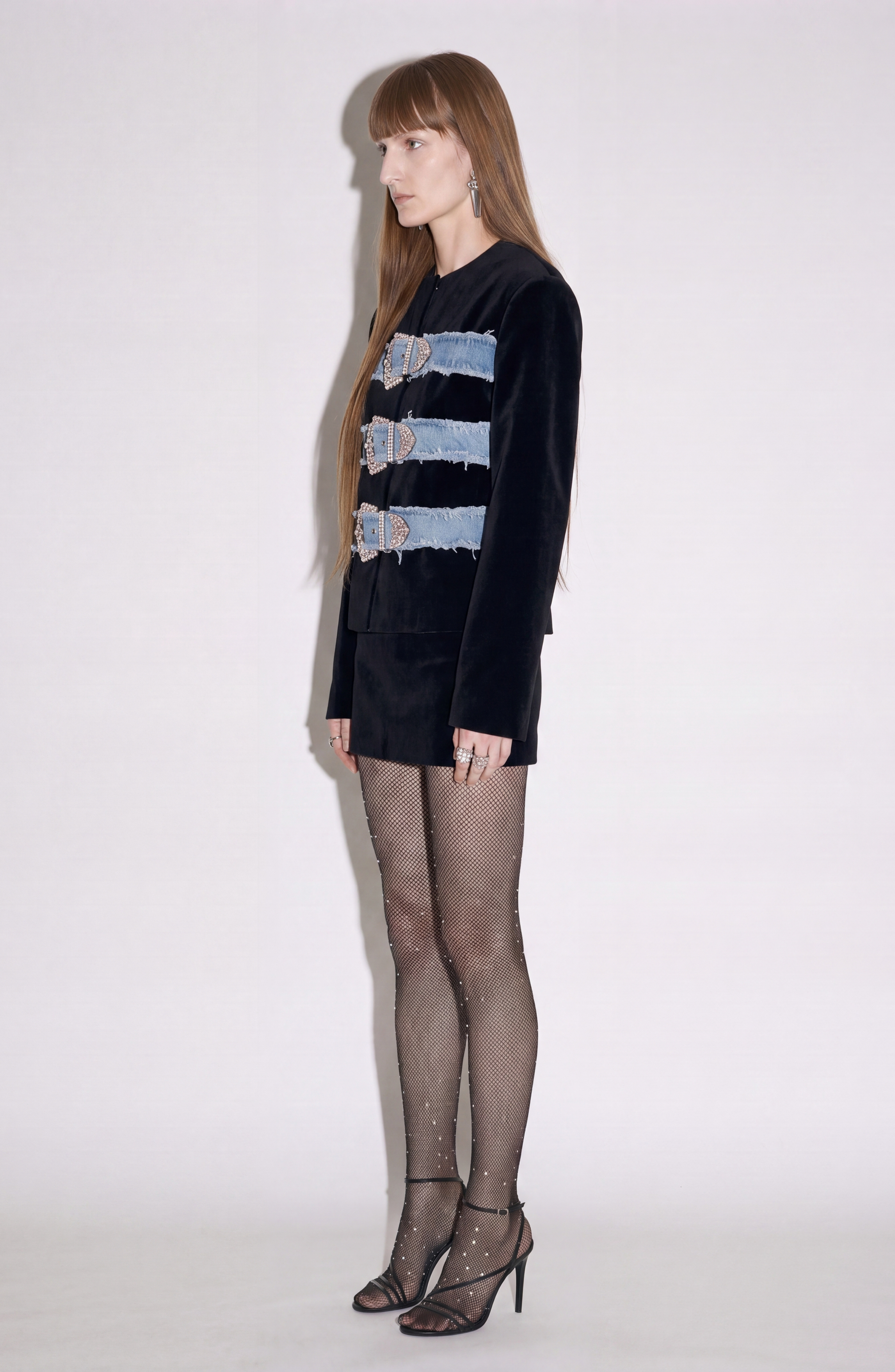 A woman with long hair stands in profile against a plain light background, wearing LOULOU’s Crystal Buckle Velvet Jacket—a collarless black velvet piece adorned with crystal-embellished buckles—paired with black shorts, sheer black rhinestone tights, and black heels.