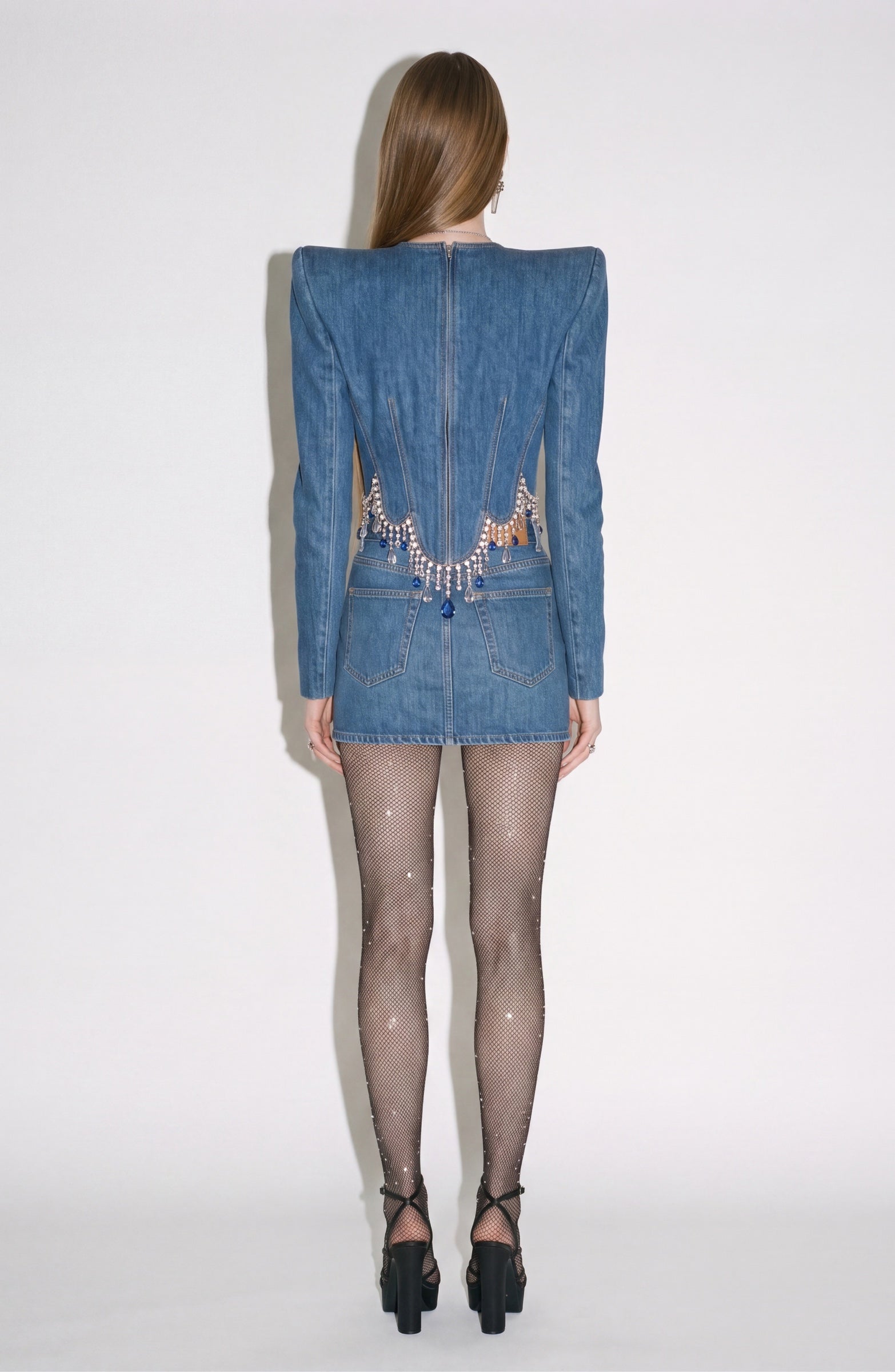A woman stands facing away, wearing the LOULOU Crystal Fringe Denim Top with sharp shoulders and a sculpted, panelled silhouette featuring rhinestone details at the waist. She pairs it with black sheer sparkly tights and black high-heeled sandals. The background is plain white.