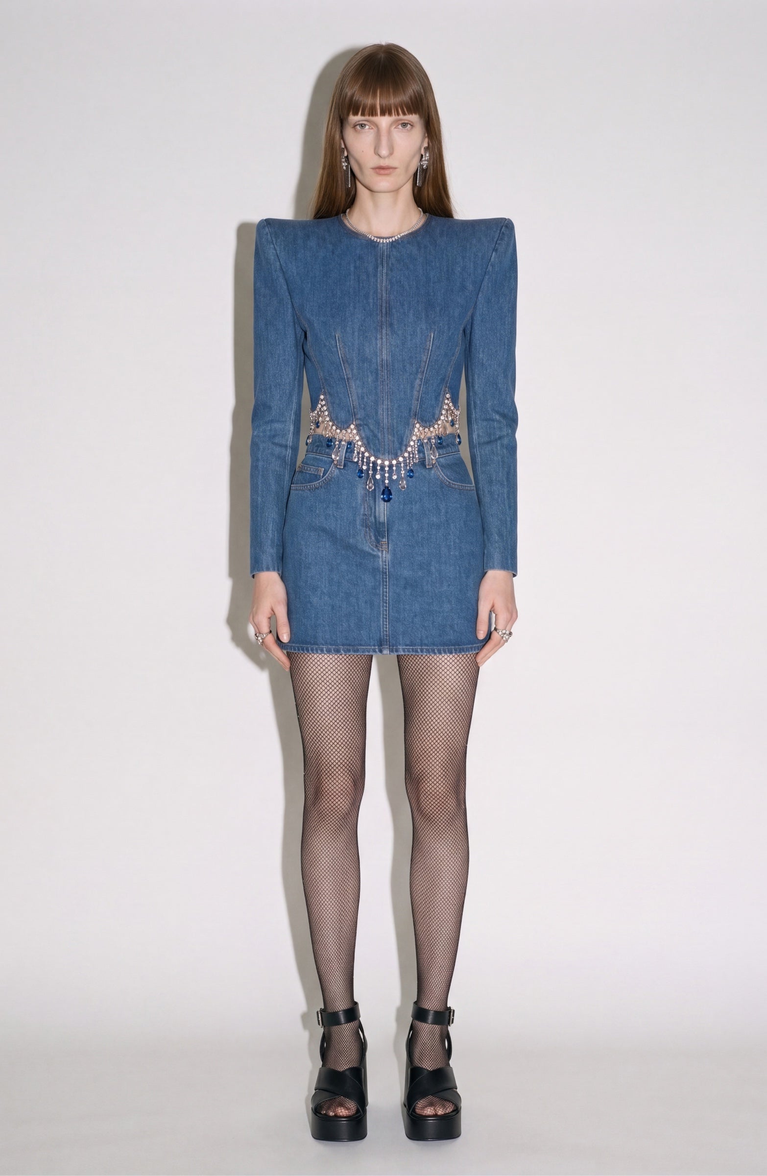 A person stands against a plain background, wearing the LOULOU Crystal Fringe Denim Top with pronounced shoulders and a sculpted, paneled body, styled with fishnet tights, black platform sandals, and a decorative belt featuring metallic details.