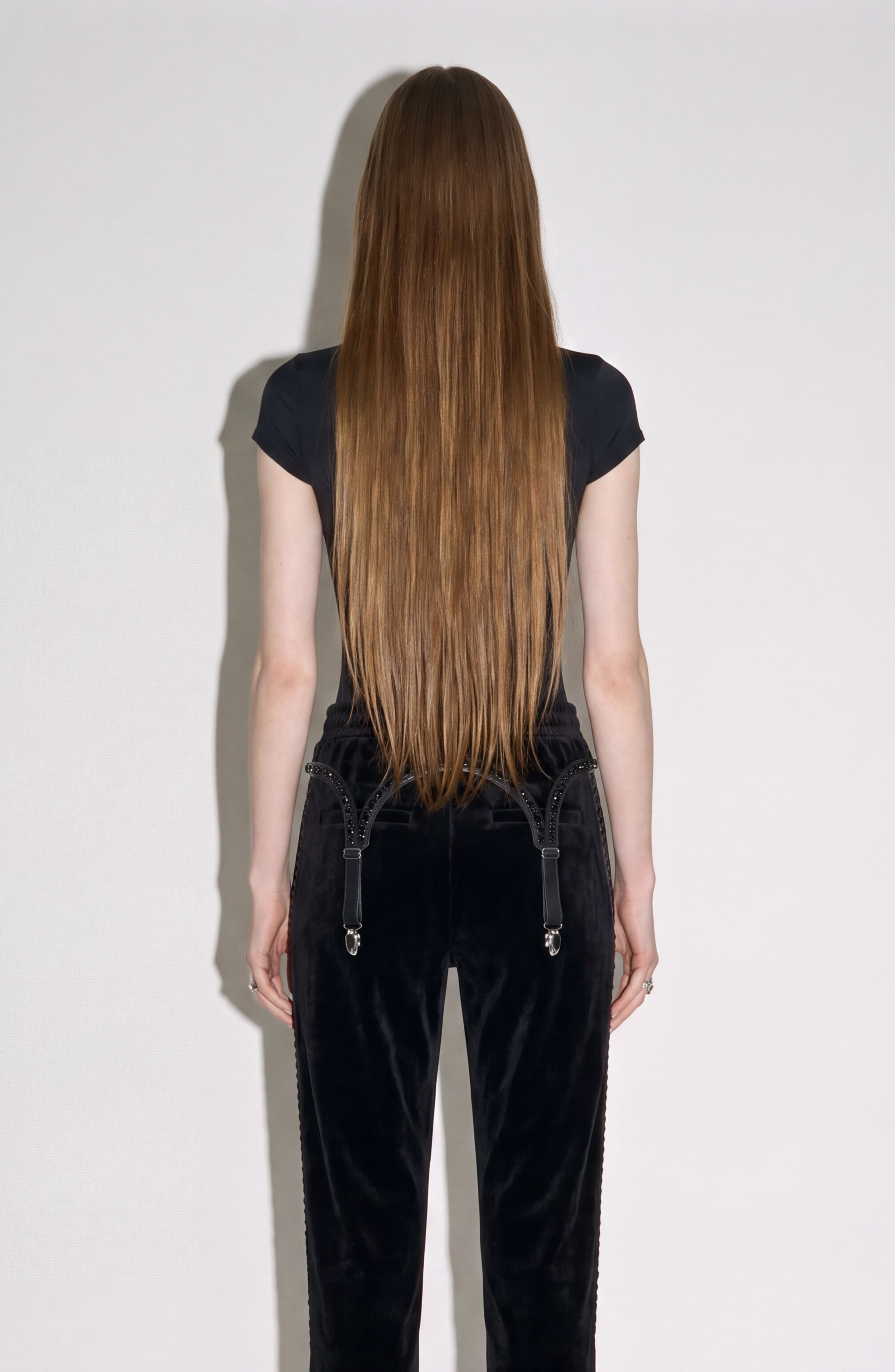 A person with very long, straight brown hair stands facing a white wall, shown from the back. They wear a black T-shirt and LOULOU’s Crystal Suspender Velvet Trousers.