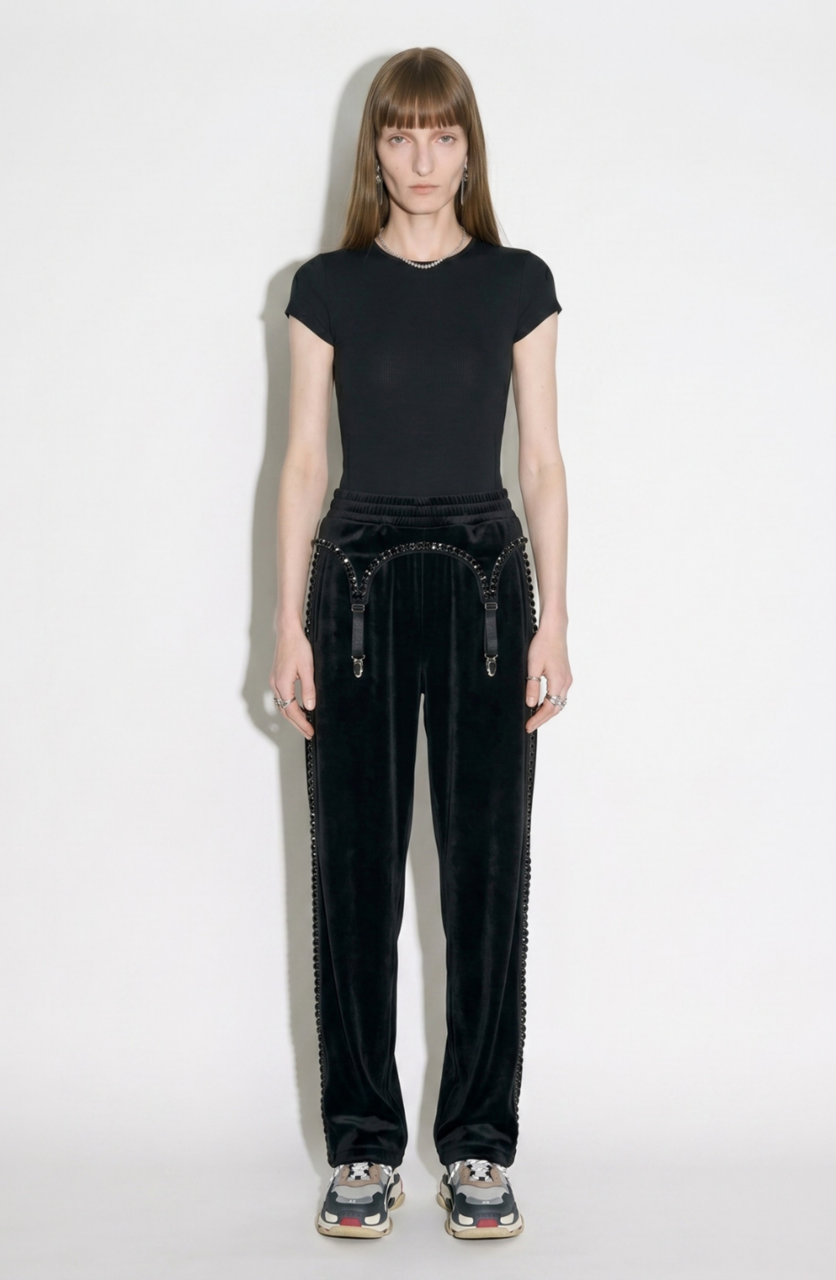 A person with straight brown hair wears a black t-shirt, LOULOU Crystal Suspender Velvet Trousers, and black sneakers, standing against a plain white background.