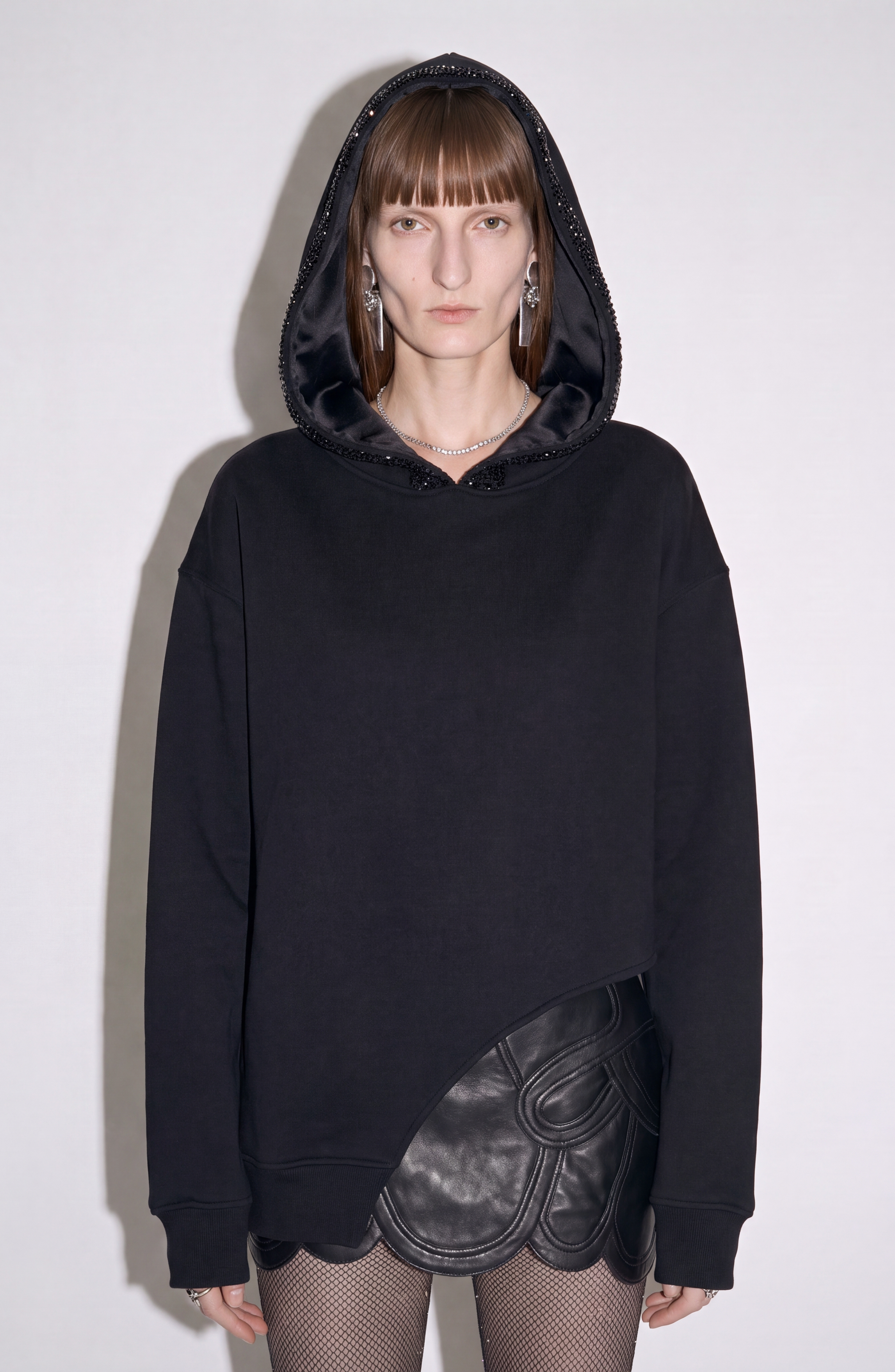 A person wearing the LOULOU Crystal Trim Asymmetrical Hoodie—an oversized black design crafted from heavyweight cotton jersey with a studded, crystal-embellished hood—paired with a black scalloped leather skirt and fishnet stockings stands against a plain, light background, looking directly at the camera.