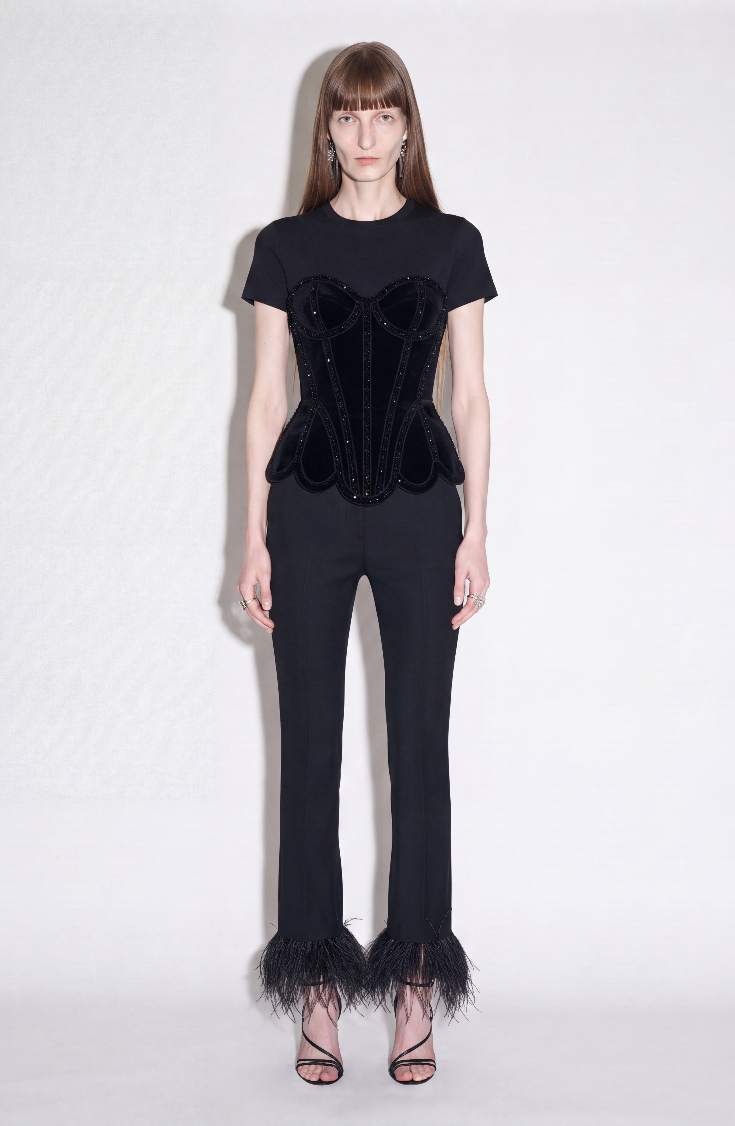 A woman stands against a plain background wearing a black cotton jersey short-sleeve top, LOULOU’s Crystal Velvet Corset T-Shirt, black pants with feathered hems, and strappy black heels.