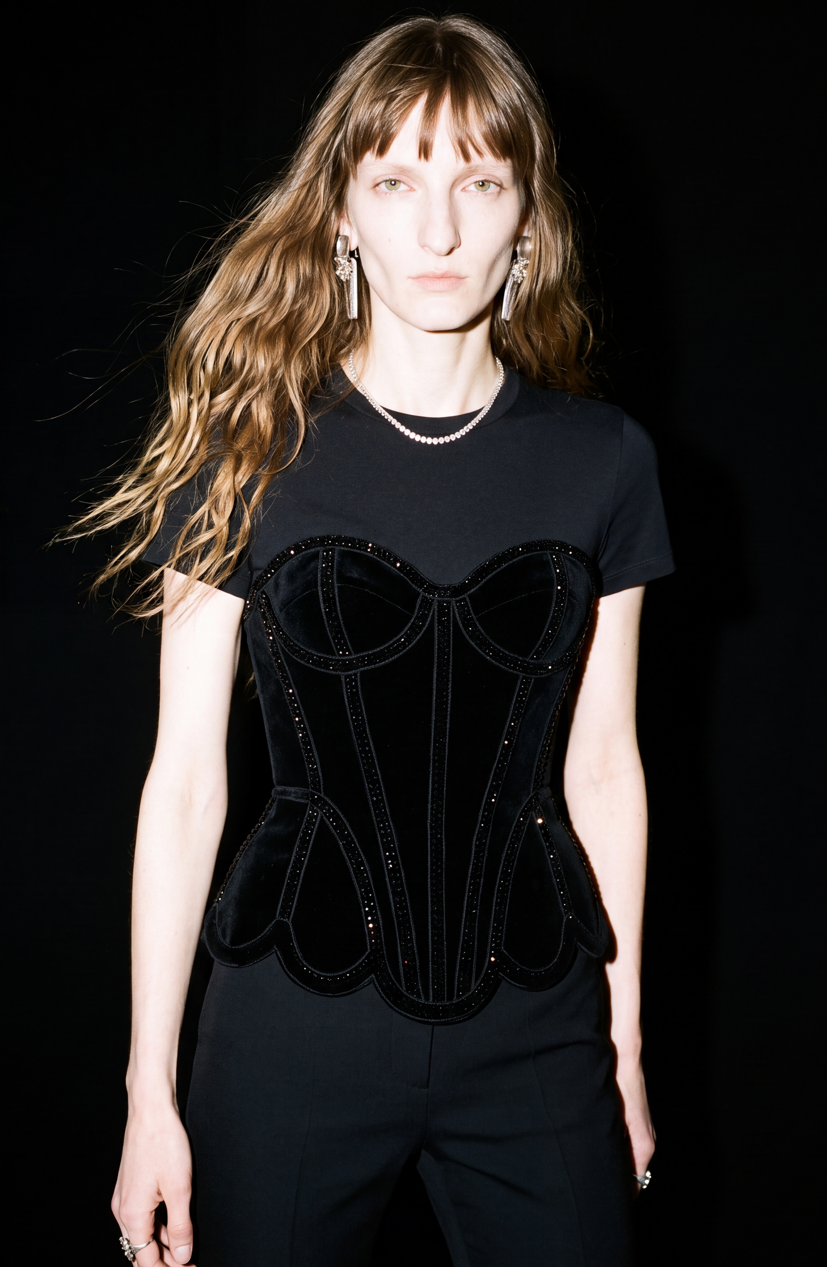 A person with long wavy hair wears a black cotton jersey T-shirt layered with the LOULOU Crystal Velvet Corset T-Shirt, featuring structured velvet and crystal embellishments, standing against a black background.