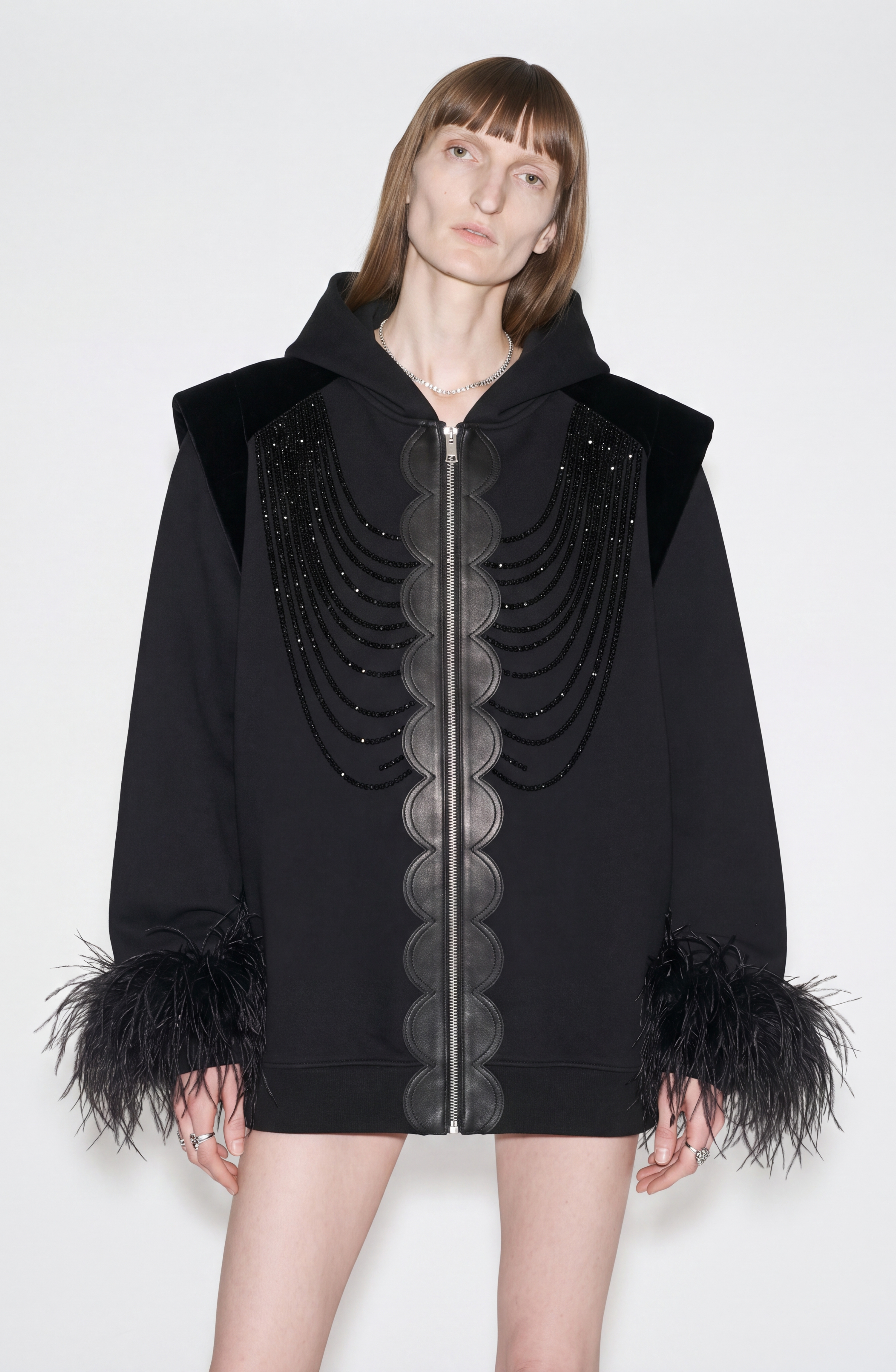 A person wearing the LOULOU Embellished Feather Trim Zip Hoodie—an oversized jet black hoodie featuring scalloped leather trim, bead embellishments, and feather-trimmed cuffs—stands against a plain white background.