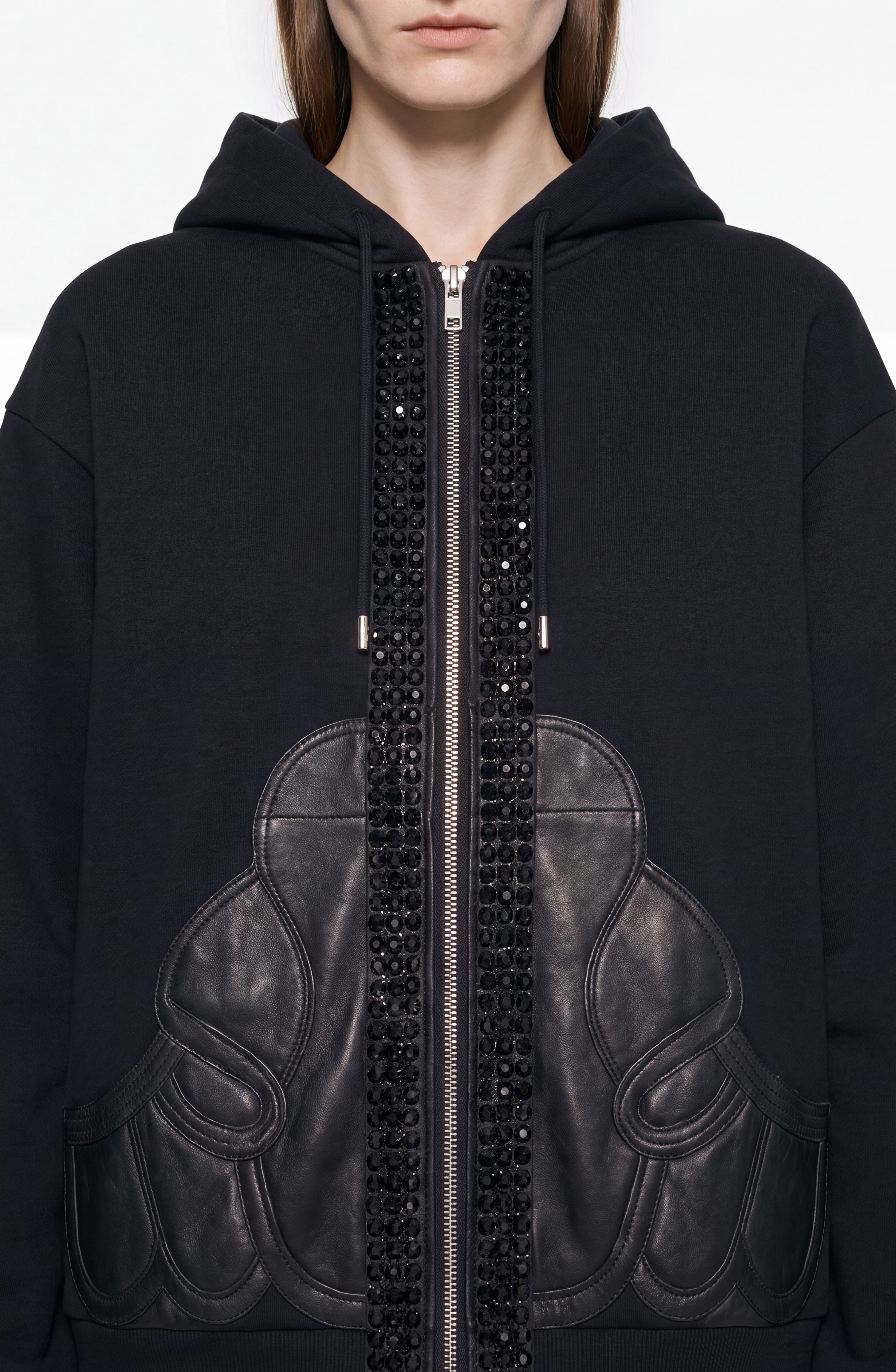 A person is shown from the lower face to upper torso, wearing the LOULOU Embellished Leather Panel Zip Hoodie in jet black. The hoodie features leather appliqué and black crystal embellishments accenting the zipper and front, set against a plain background.