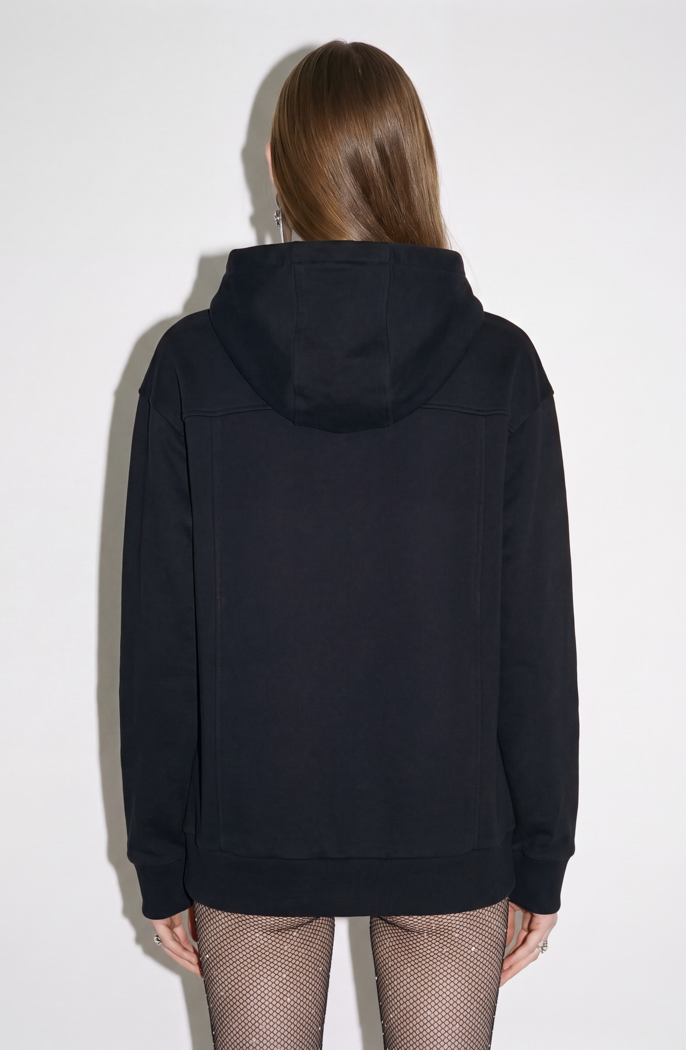 A person with long, straight brown hair stands with their back to the camera against a plain white background, wearing the LOULOU Embellished Leather Panel Zip Hoodie and fishnet tights.