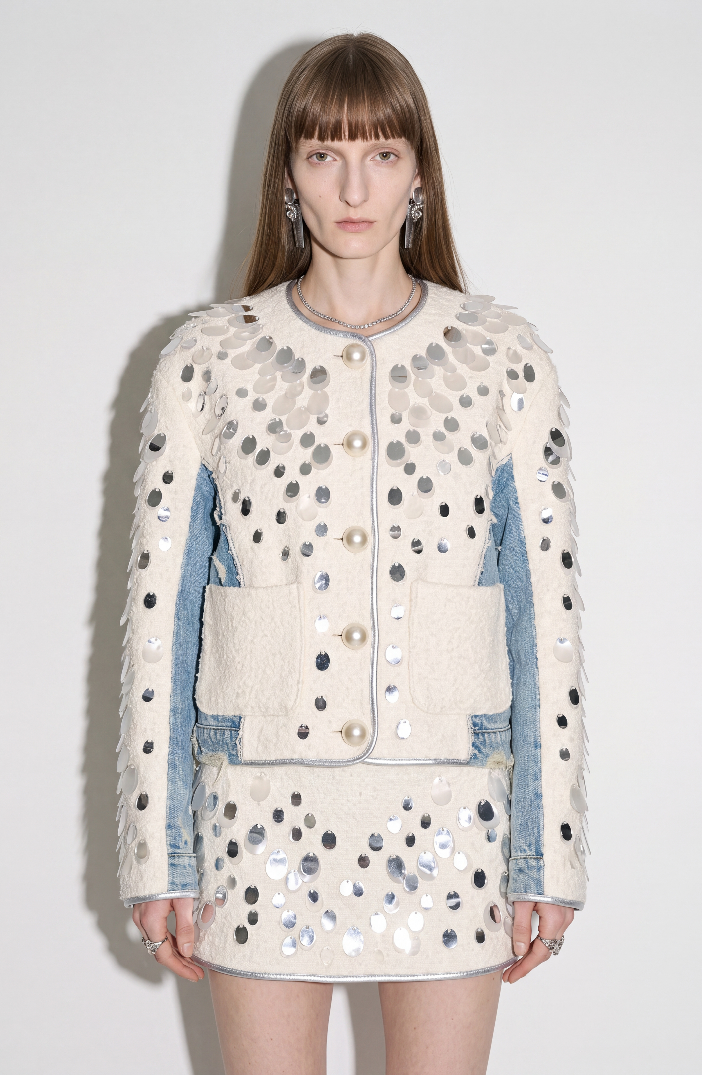 A model stands against a plain white background, looking directly at the camera while wearing the LOULOU Embellished Panelled Cropped Jacket and matching skirt, both featuring large silver sequins. The cream and light blue ensemble is finished with ivory textured panels and embellished pearl buttons for an elegant touch.