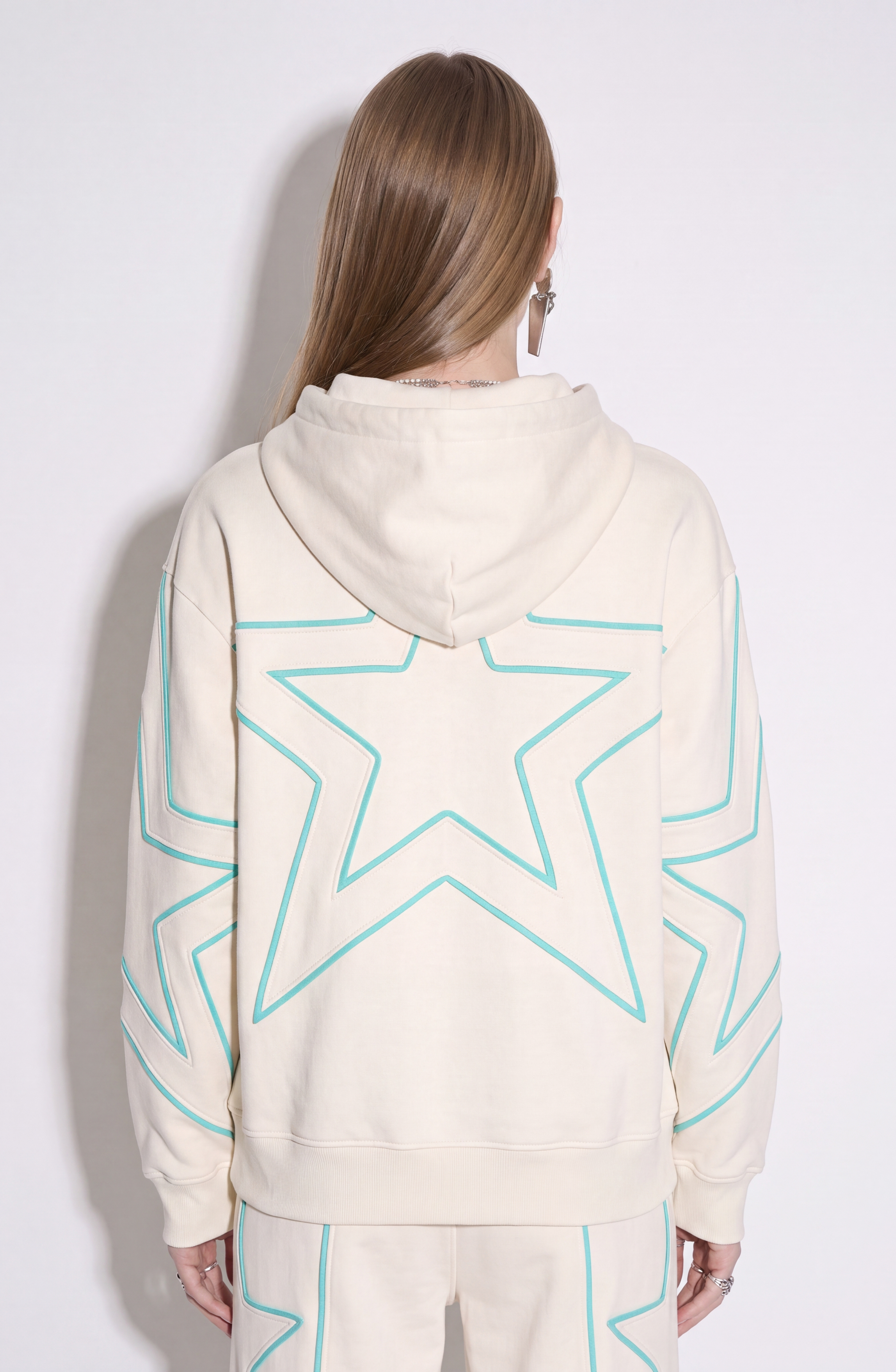 A person with long, straight hair is seen from behind against a plain white background, wearing the LOULOU Embroidered Star Cotton Hoodie— a cream cotton jersey piece decorated with large outlined turquoise stars on the back and sleeves—paired with matching pants.