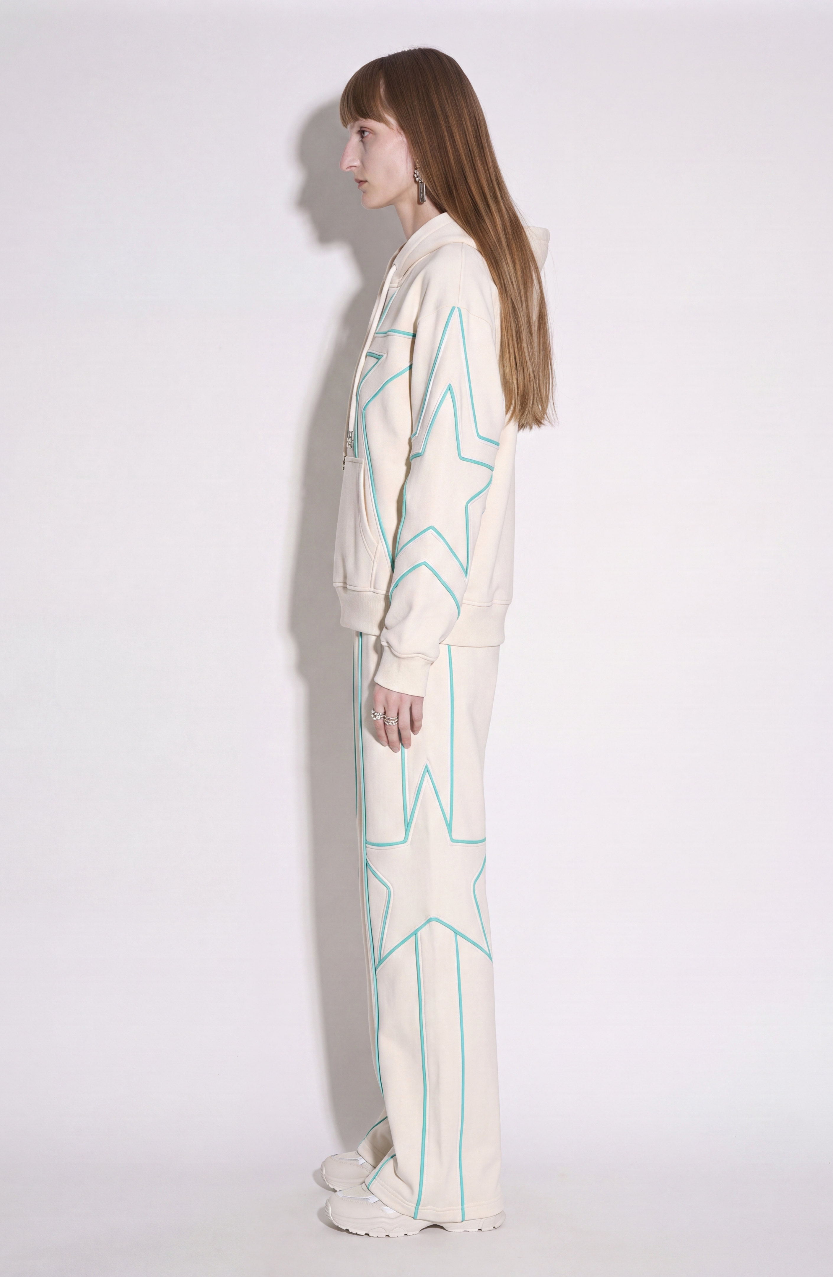 A person with long hair stands sideways against a plain white background, wearing the LOULOU Embroidered Star Cotton Hoodie—a cream-colored cotton jersey tracksuit adorned with large light blue and turquoise star designs on both the jacket and pants, featuring a cozy hoodie.