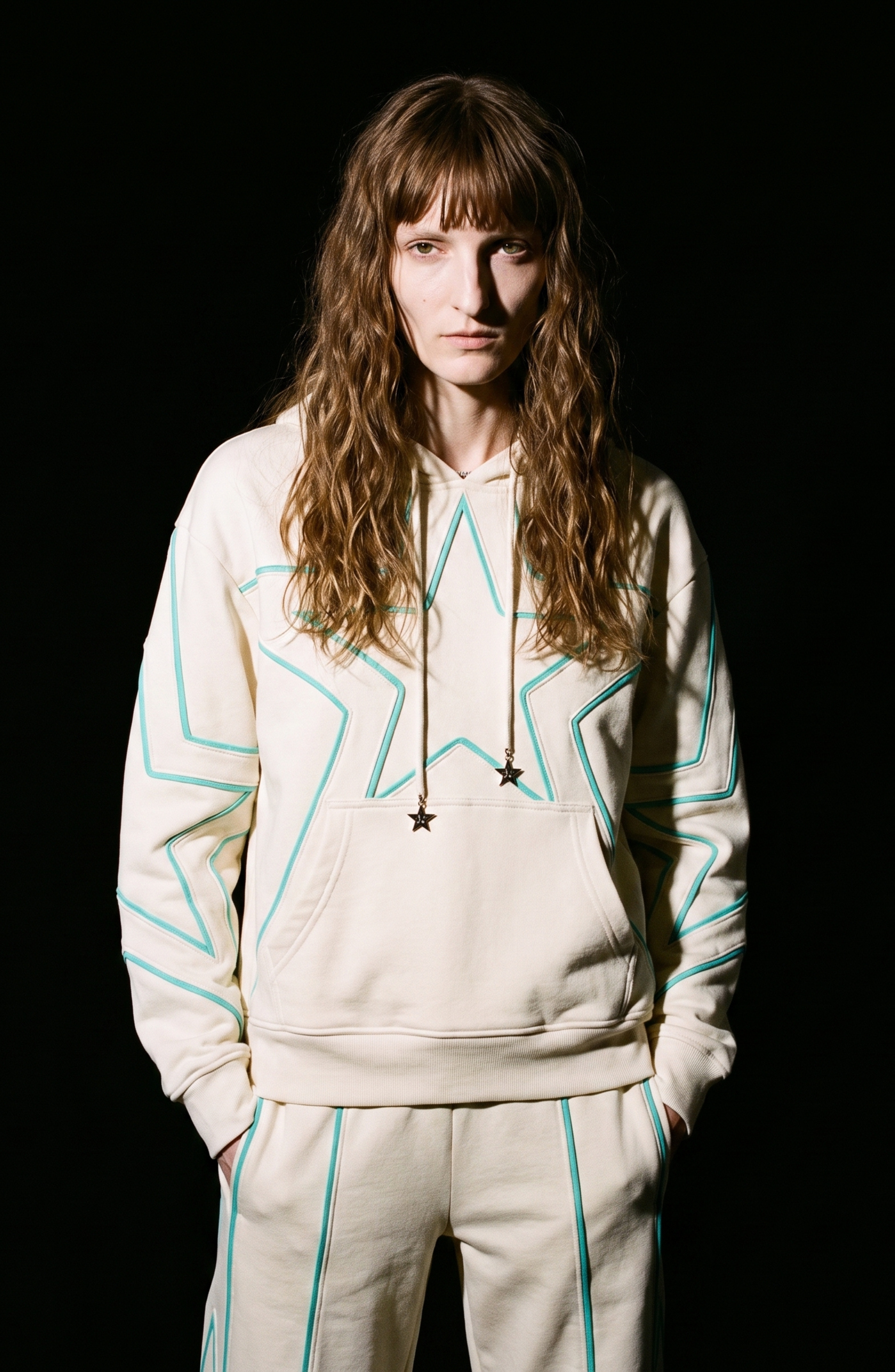 A person with long, wavy hair models the LOULOU Embroidered Star Sweatpants and a coordinating cream cotton jersey hoodie, both adorned with turquoise star embroidery, while standing against a black background and gazing directly at the camera.