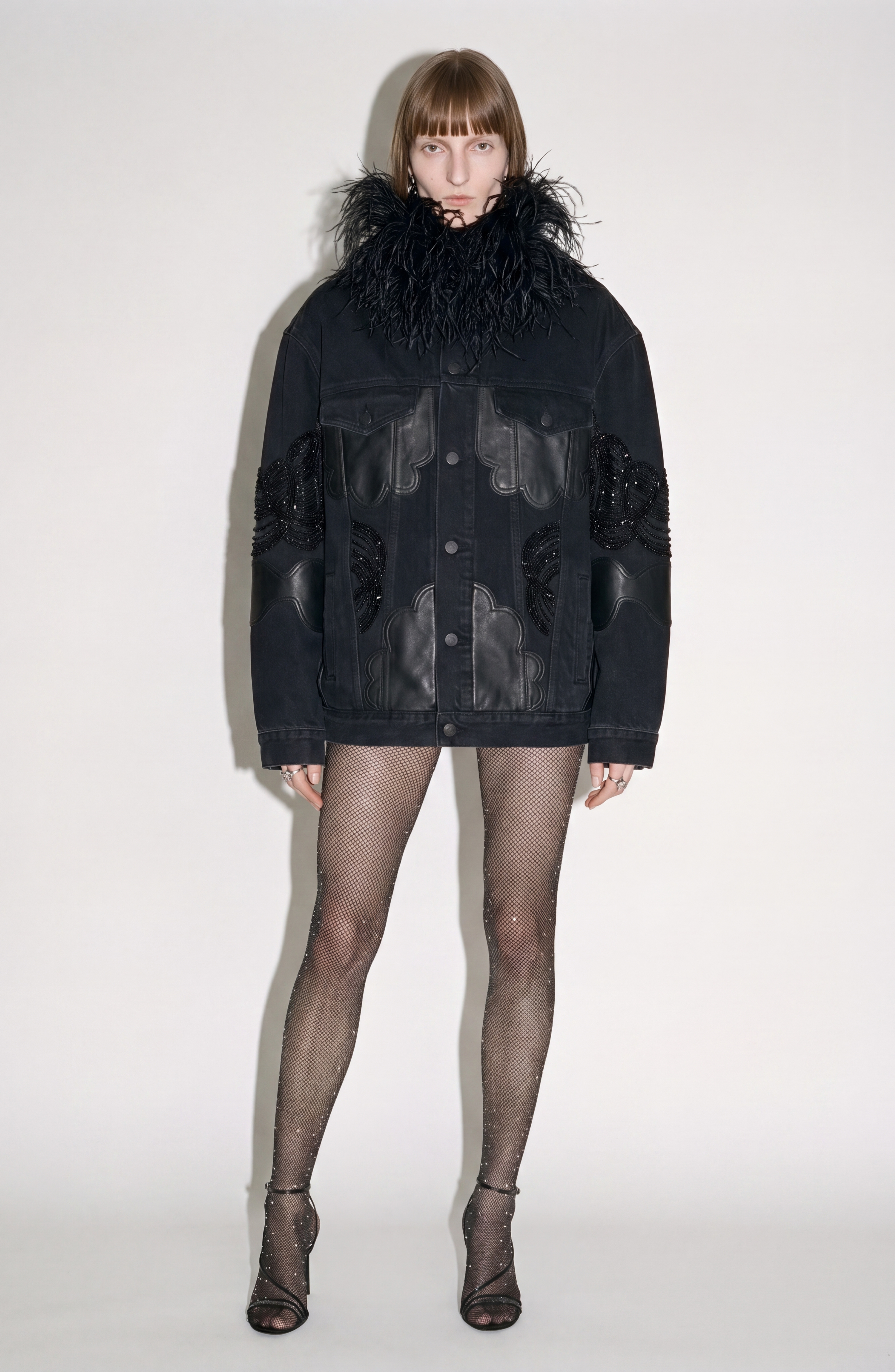 A person stands against a plain white background wearing the LOULOU Feather-Trimmed Appliqué Denim Jacket, paired with sheer sparkly tights and black high-heeled sandals.