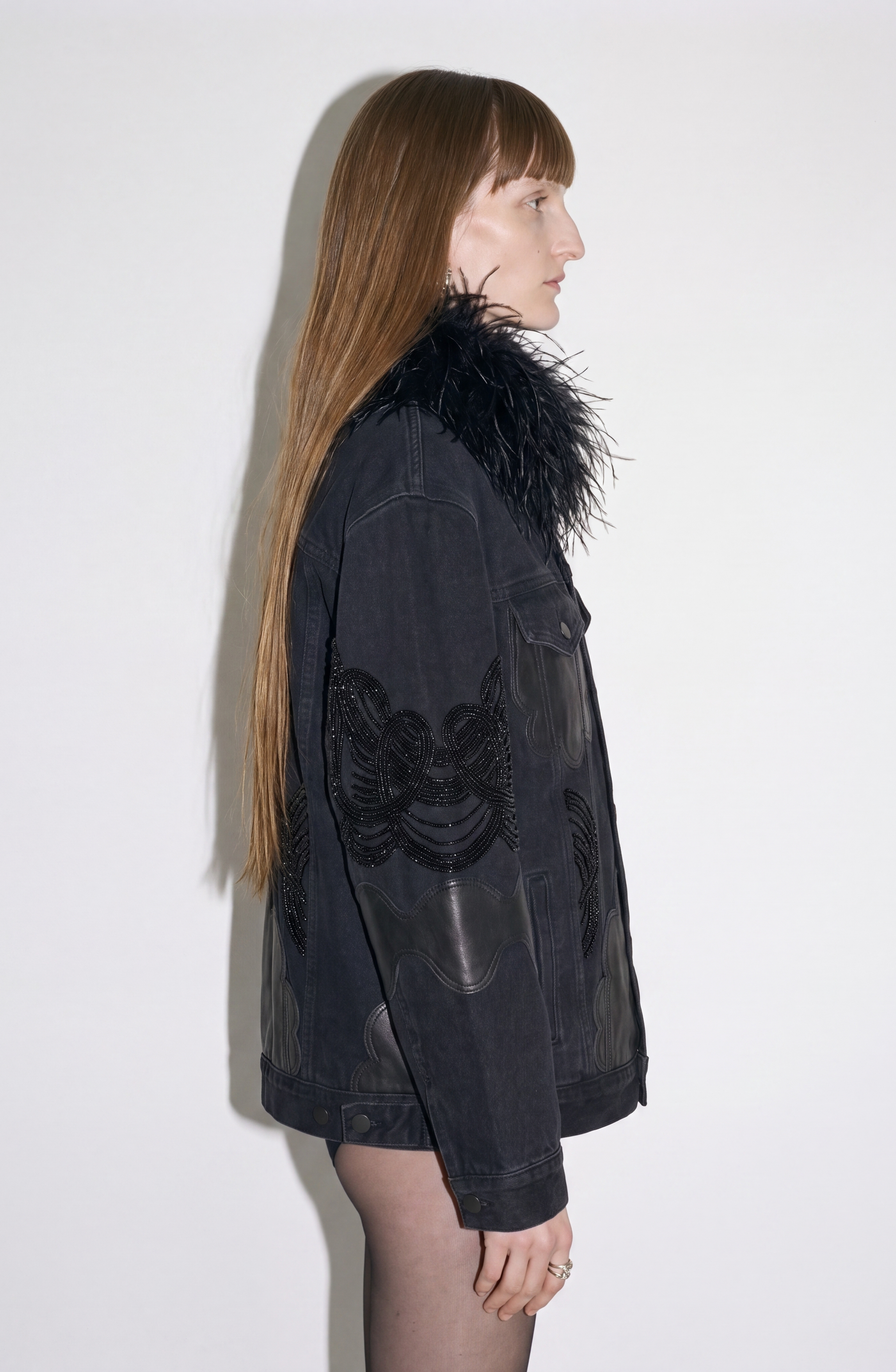 A person with long straight hair stands in profile against a plain white background, wearing the LOULOU Feather-Trimmed Appliqué Denim Jacket—an oversized piece featuring a feather collar and intricate leather appliqué detailing.