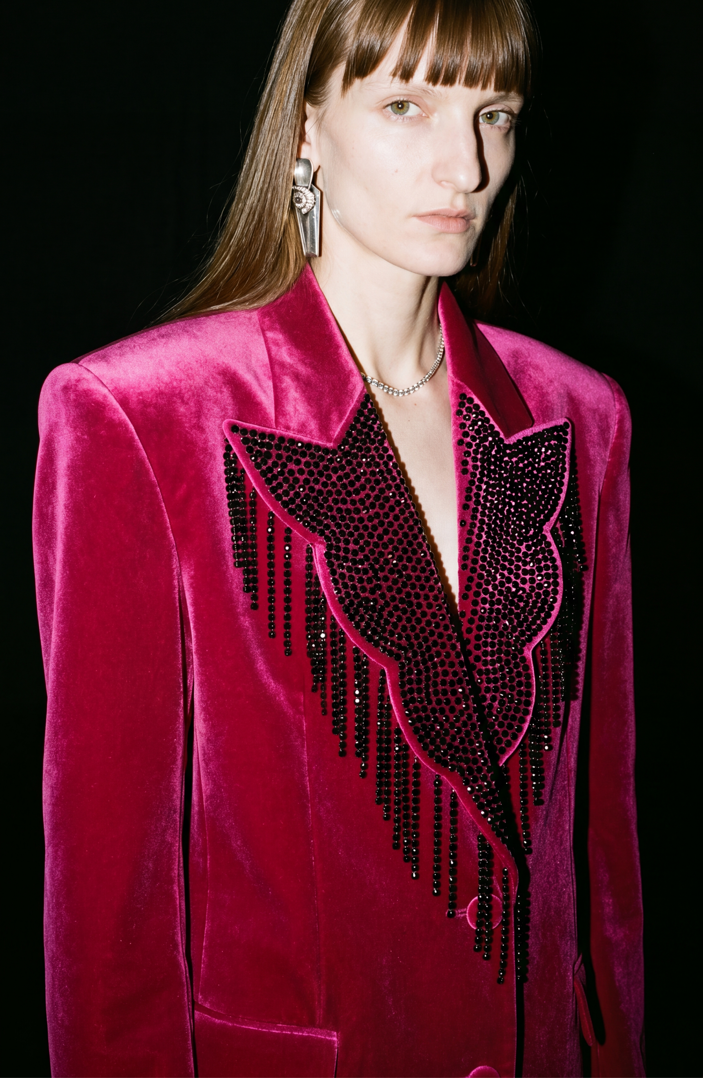 A person with long brown hair wears the LOULOU Fuchsia Velvet Blazer with black beaded fringe and geometric designs, paired with silver earrings and a necklace, standing against a black background.