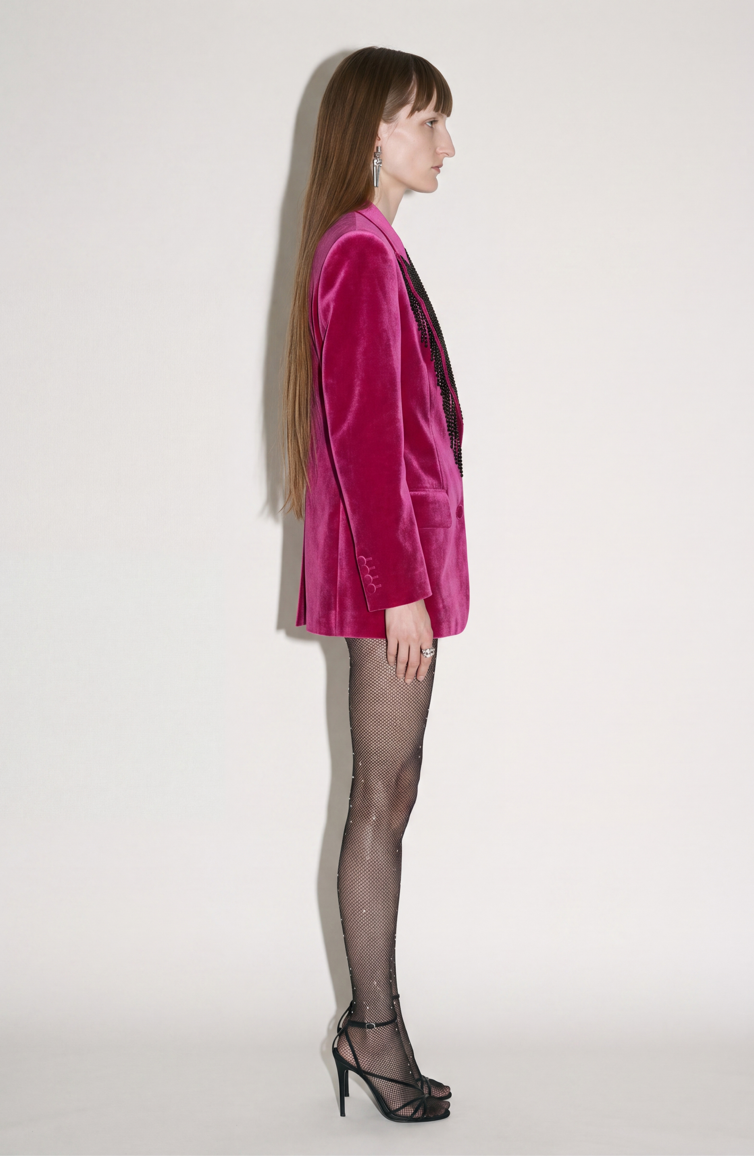 A person stands in profile against a plain background, wearing LOULOU’s Fuchsia Velvet Blazer, sheer black embellished tights, black high heels, and statement earrings. Their long straight brown hair completes the look.