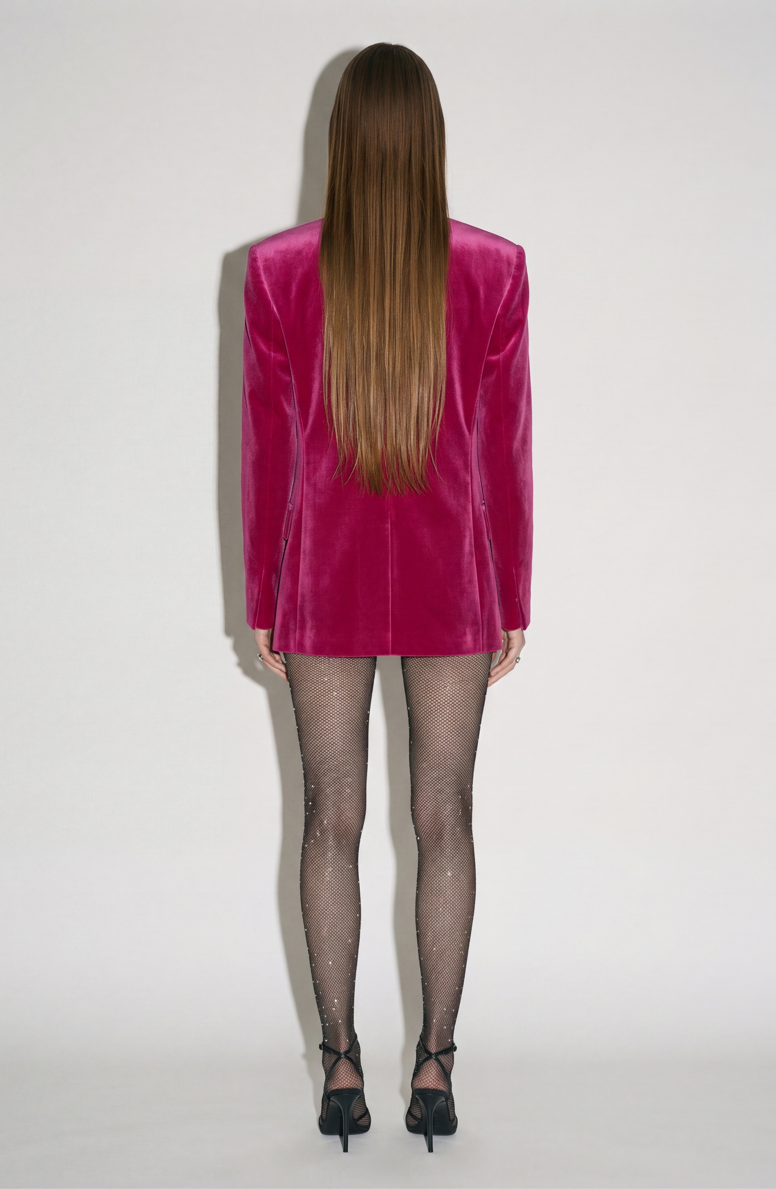 A person with long straight hair faces away, wearing the LOULOU Fuchsia Velvet Blazer, sparkling black sheer tights, and black heels against a plain light background.