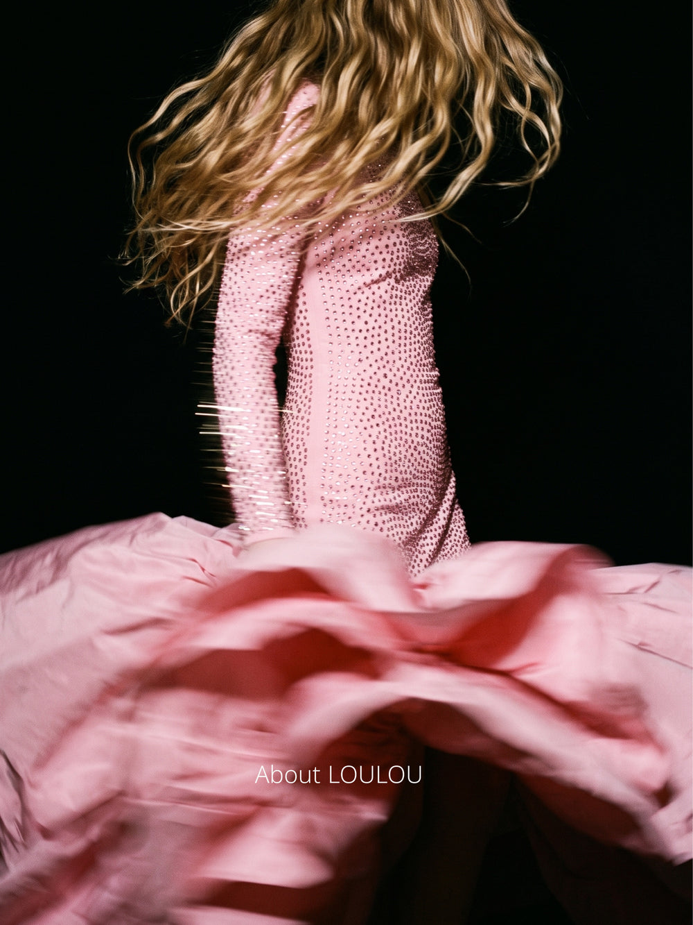 Model wearing a blush pink crystal embellished gown from the LOULOU collection captured in motion against a black backdrop.