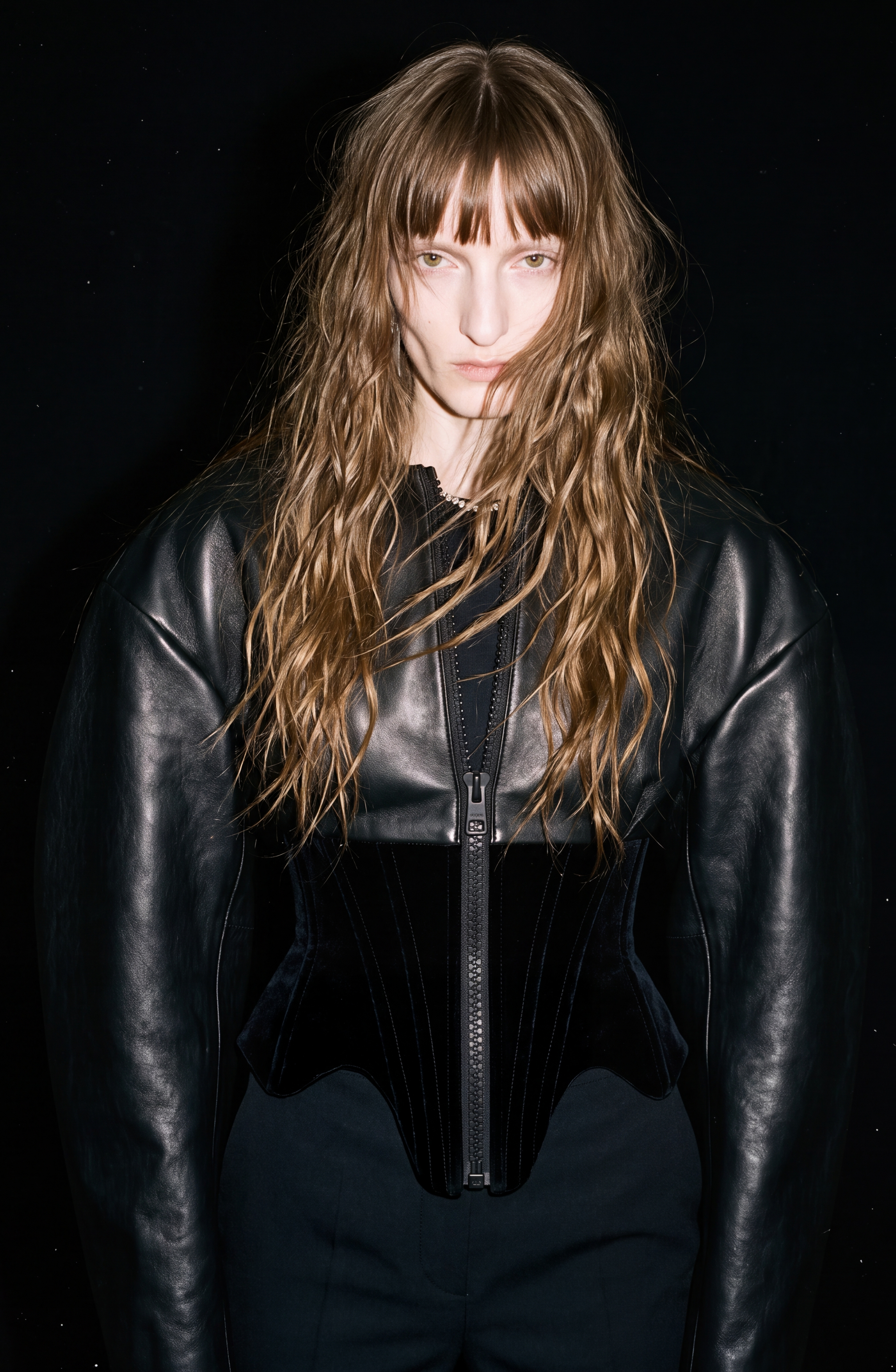 A person with long, wavy hair wears the LOULOU Leather And Velvet Corset Jacket, featuring voluminous sleeves and a structured silhouette. They stand against a dark background, looking directly at the camera with a neutral expression.
