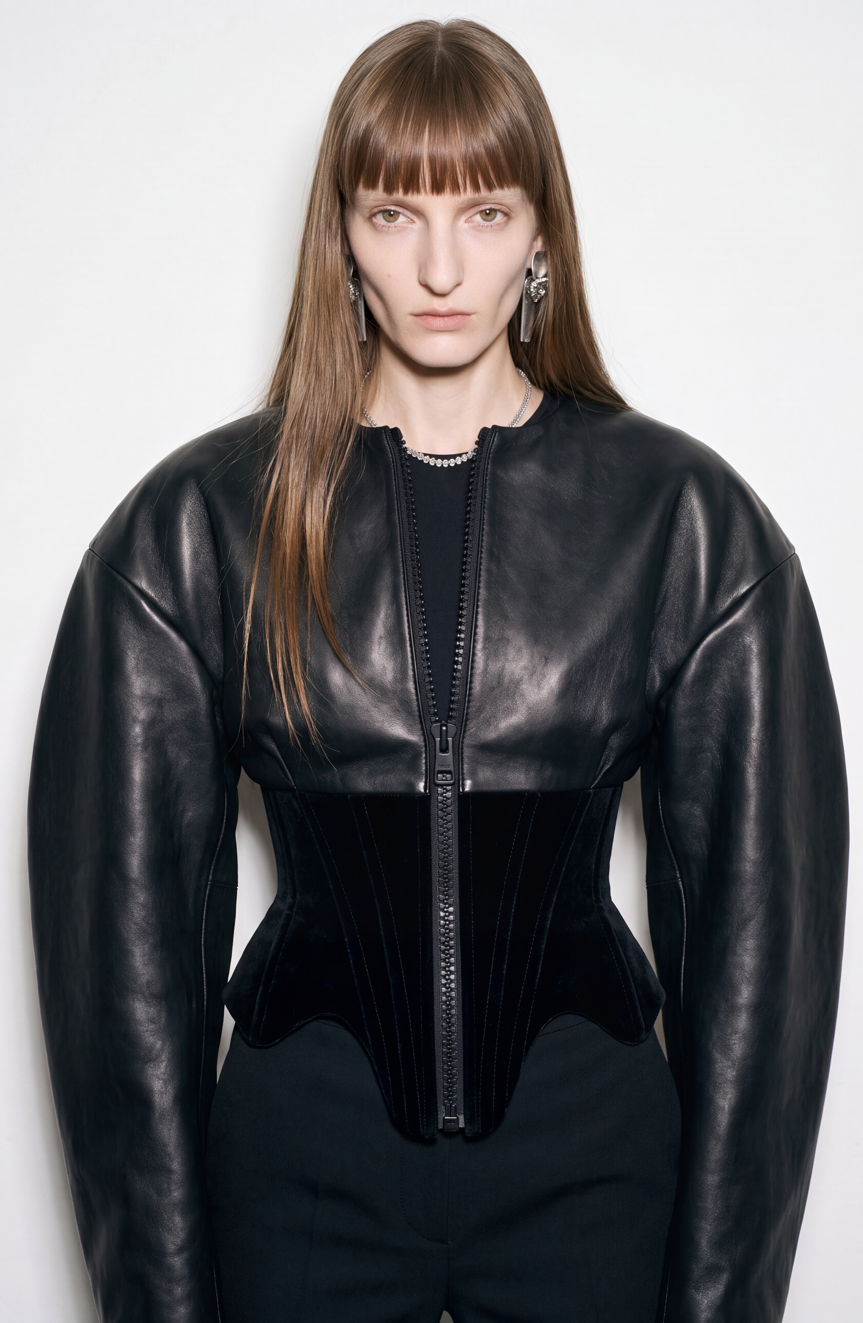 A person with long straight hair and blunt bangs wears the LOULOU Leather And Velvet Corset Jacket, featuring voluminous sleeves, paired with black pants and statement earrings, standing against a white background.