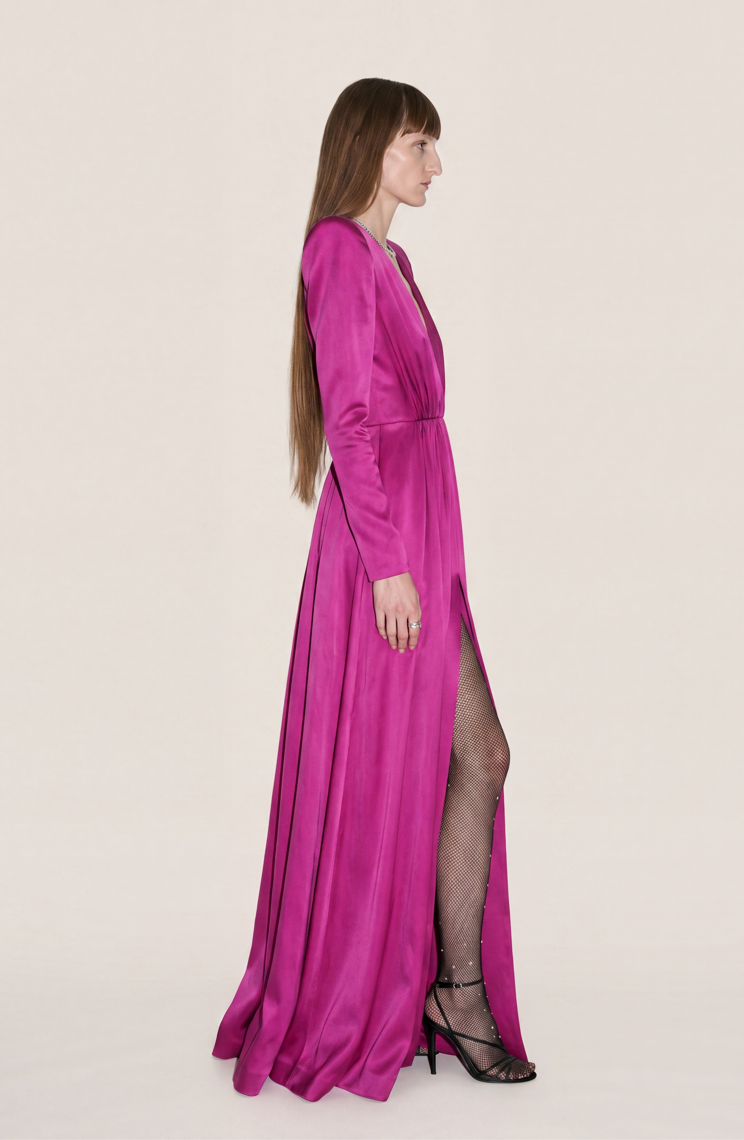 A woman with long brown hair stands in profile against a plain light background, wearing the LOULOU Magenta Satin Gown—a full-length dress featuring a high slit, paired with fishnet stockings and black heels.