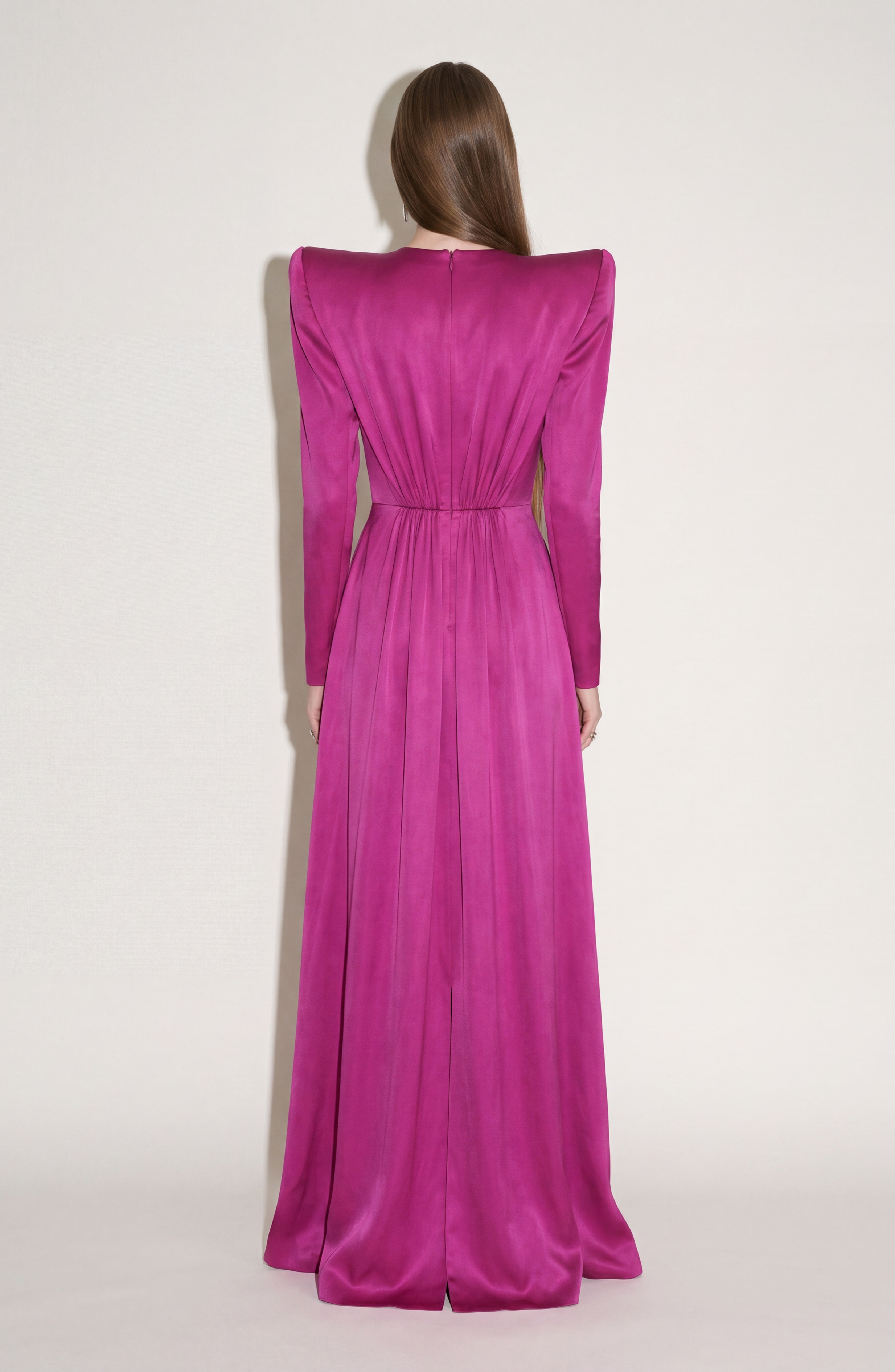 A person with long brown hair faces away, wearing the LOULOU Magenta Satin Gown with a full-length silhouette, defined waist, and structured shoulders, set against a plain light background.