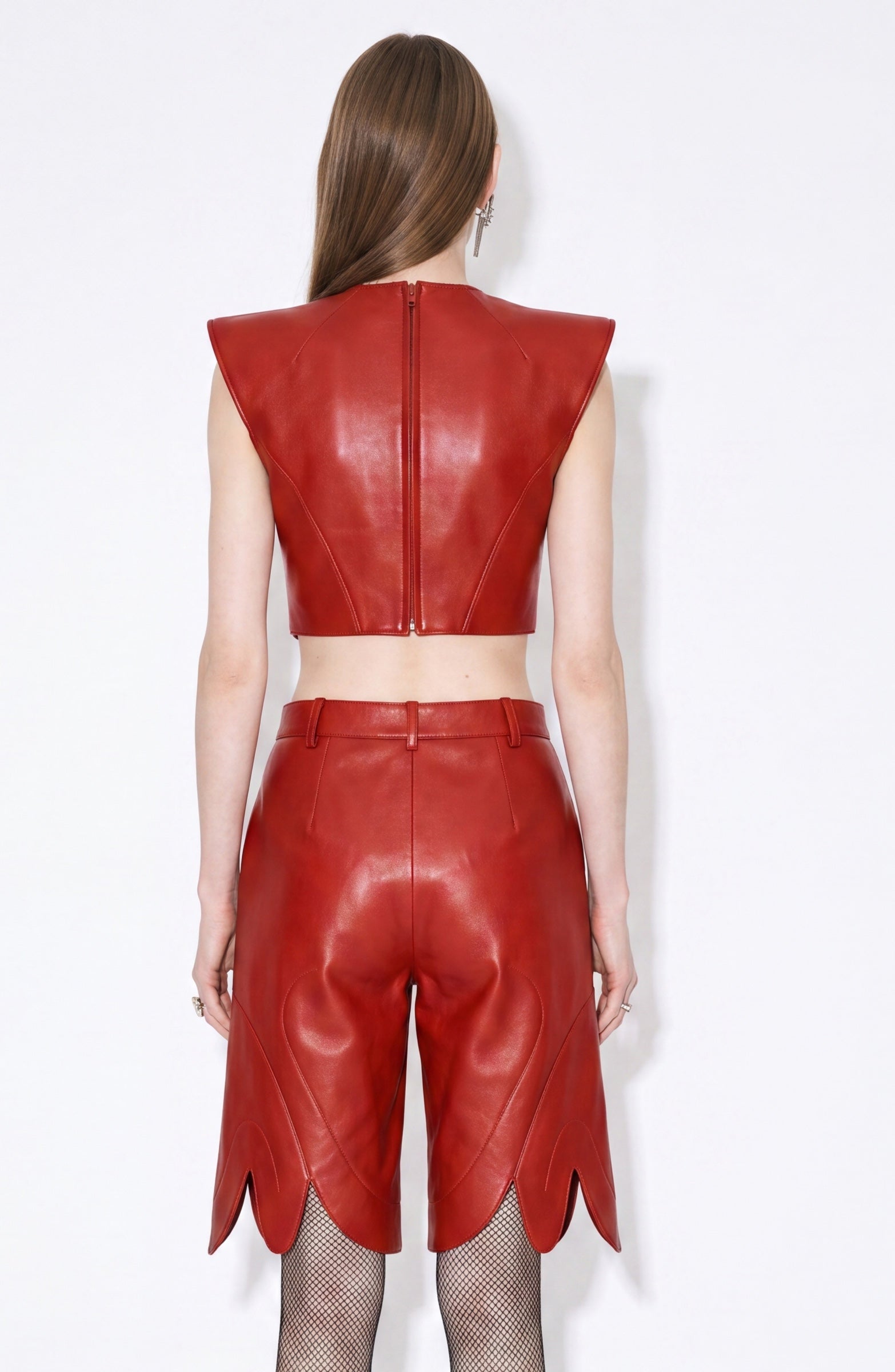 A person stands with their back to the camera, dressed in a sleeveless red leather crop top and matching LOULOU Red Leather Bermuda Shorts, featuring high-waisted lamb leather construction and scalloped hems, paired with fishnet stockings.
