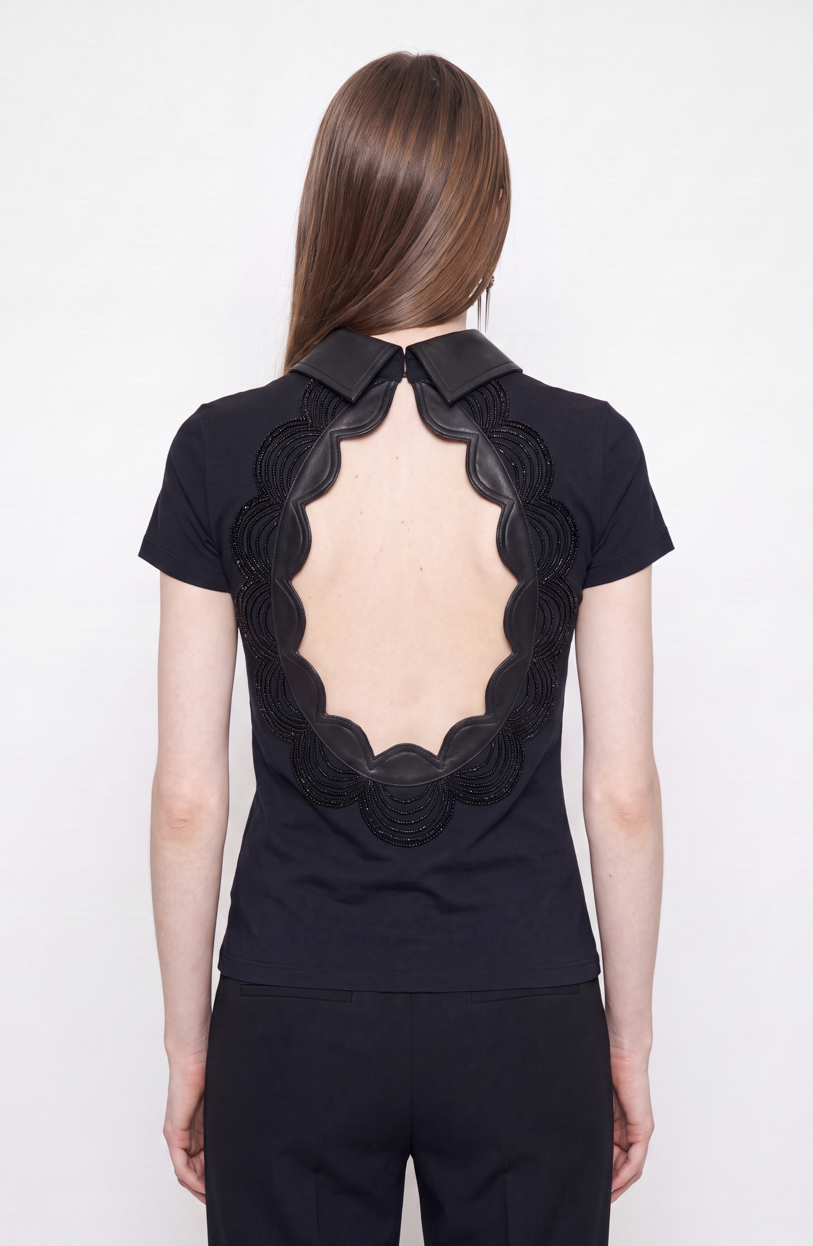 A person with long straight hair wears the LOULOU Scalloped Open Back Top— a black cotton blend fitted top with a large oval-shaped open back trimmed in scalloped leather—paired with black pants, standing against a plain light background.