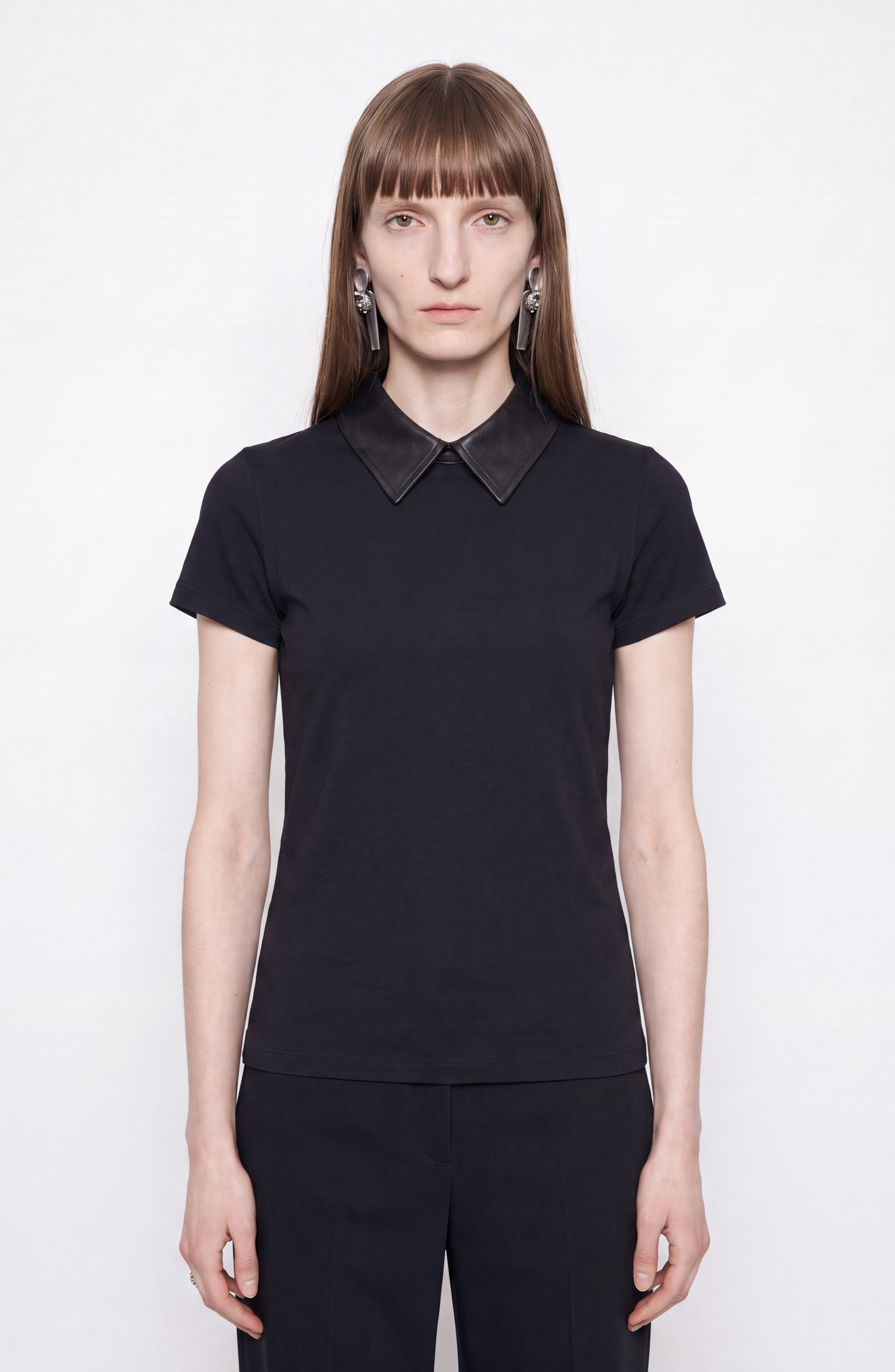 A woman with straight brown hair and bangs wears the LOULOU Scalloped Open Back Top—a black cotton blend fitted style with a leather collar—and statement earrings, standing against a plain white background.